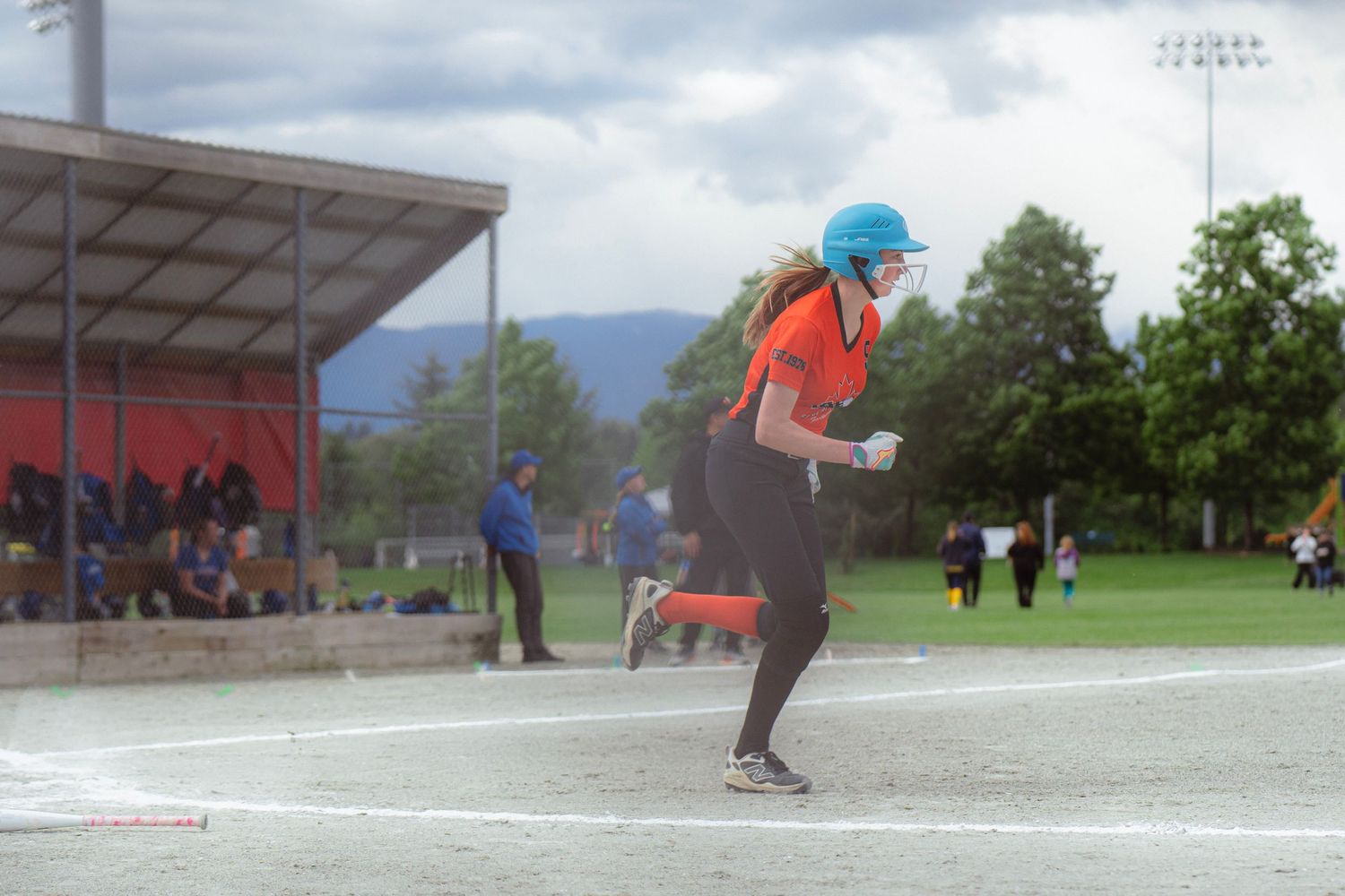 Female softball player in orange uniform running bases on a cloudy day.