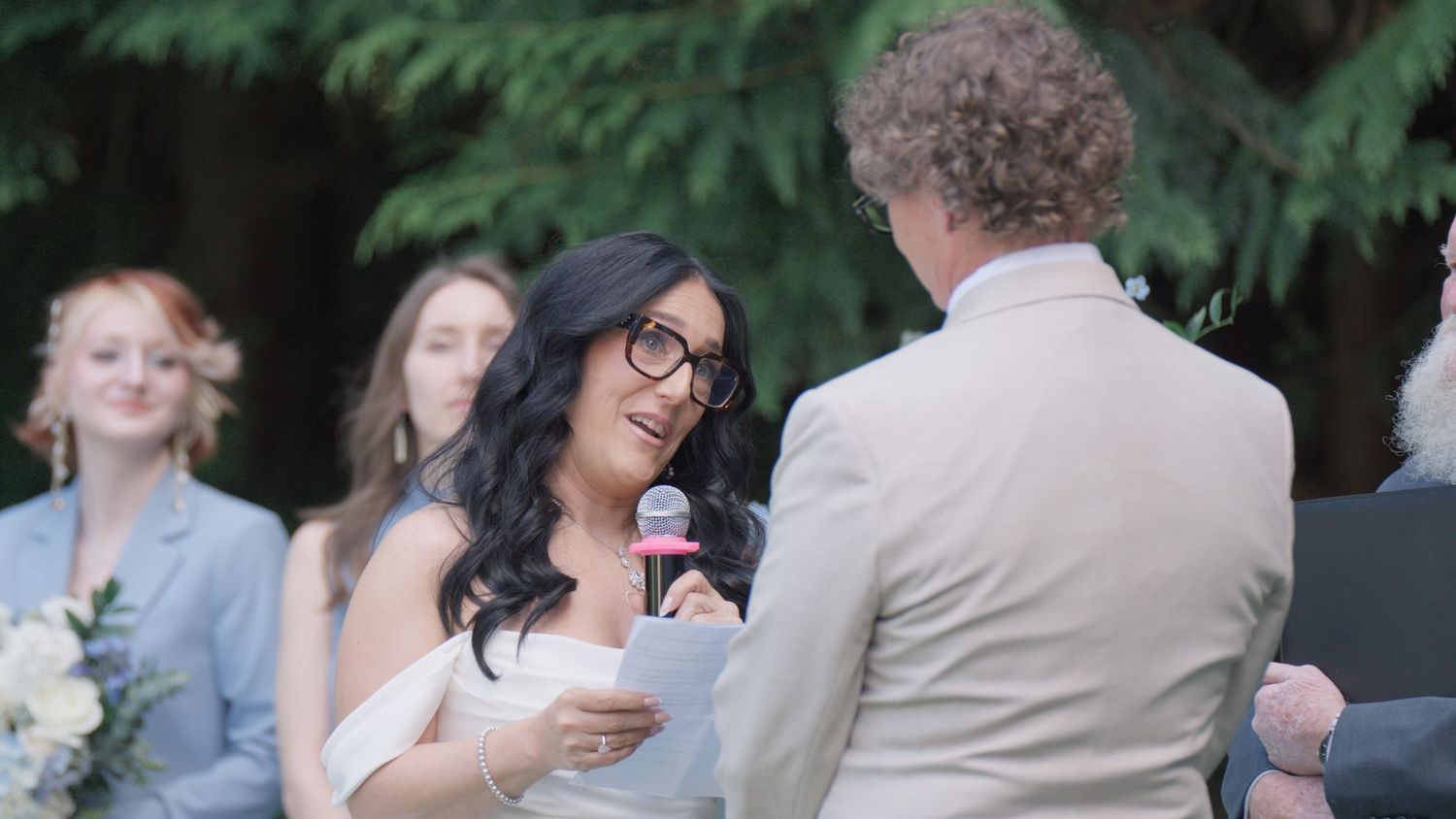 Bride in white dress reads vows at outdoor wedding ceremony while groom listens attentively.