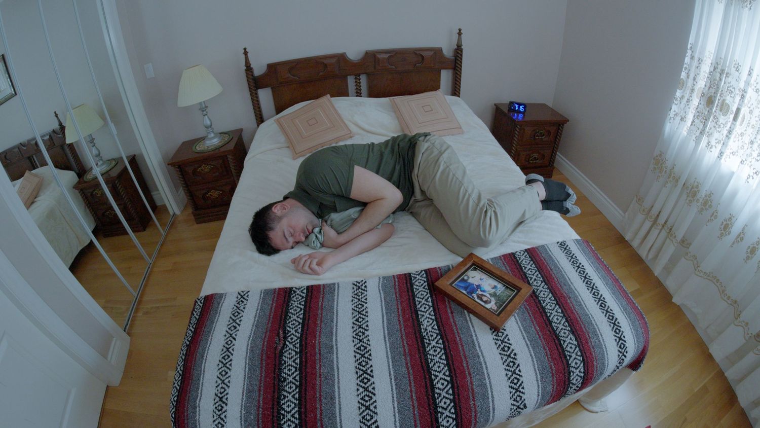 Man lying face down on a bed with a striped blanket beside a framed photo in a bright bedroom.