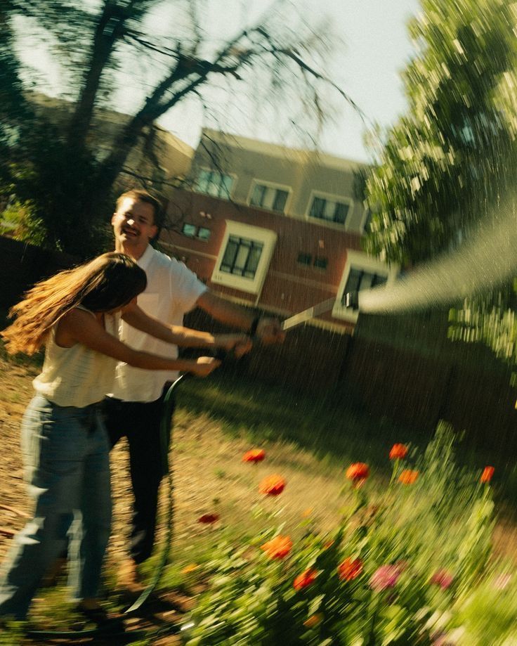 Couple laughing and spraying water with a garden hose among colorful tulips in a sunny backyard.