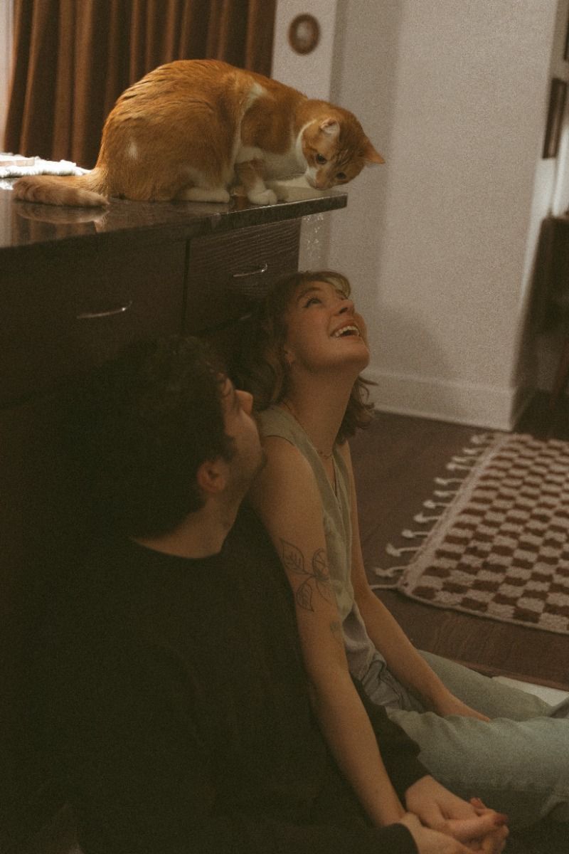 Two people sit on the floor laughing with an orange tabby cat perched on a dark dresser above them indoors.