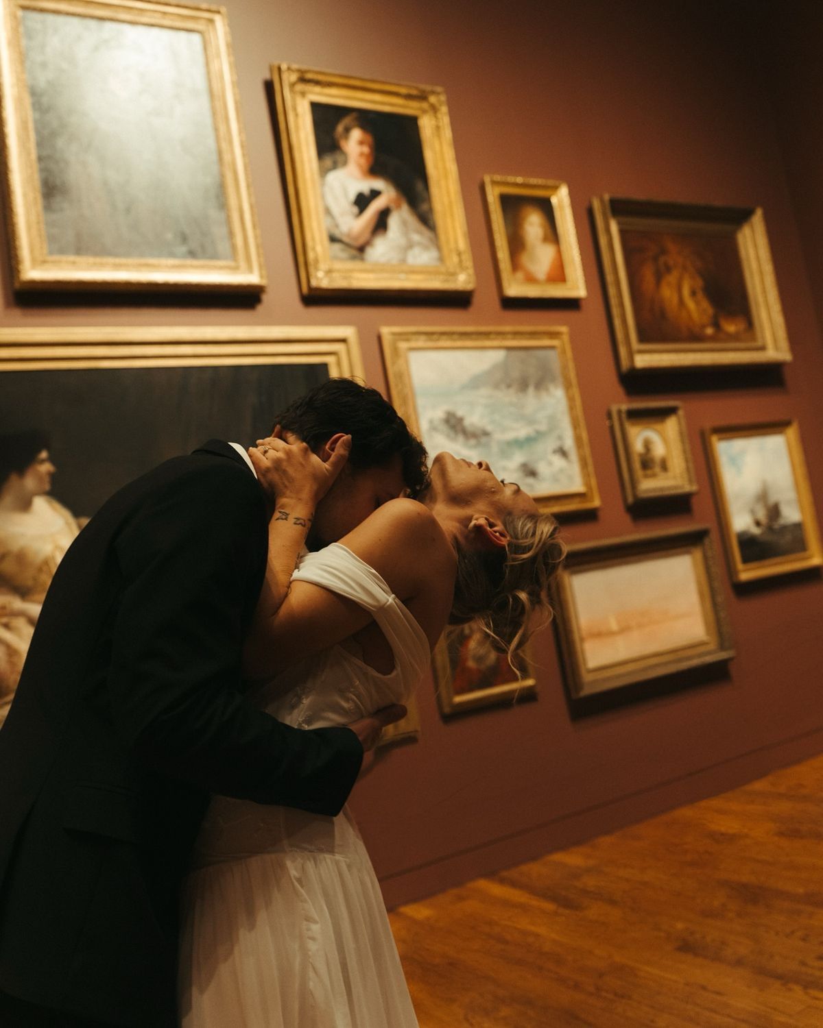 Couple sharing a romantic kiss in an art museum gallery surrounded by classical portrait paintings on warm terracotta walls.