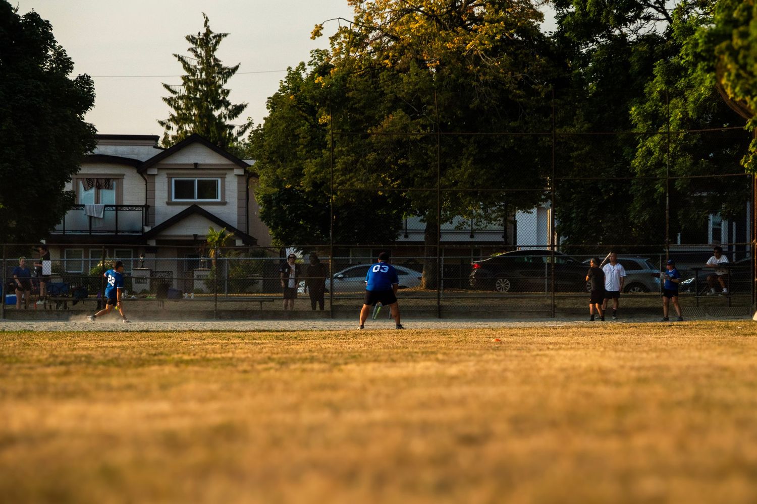 Players compete in a recreational softball game on a dusty field at golden hour with houses and trees in the background.
