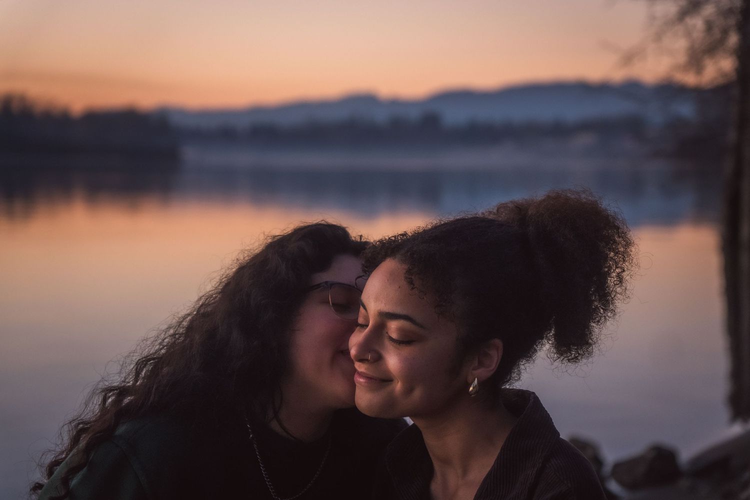 Two women share a gentle kiss on the cheek by a serene lake during a beautiful pink and orange sunset.