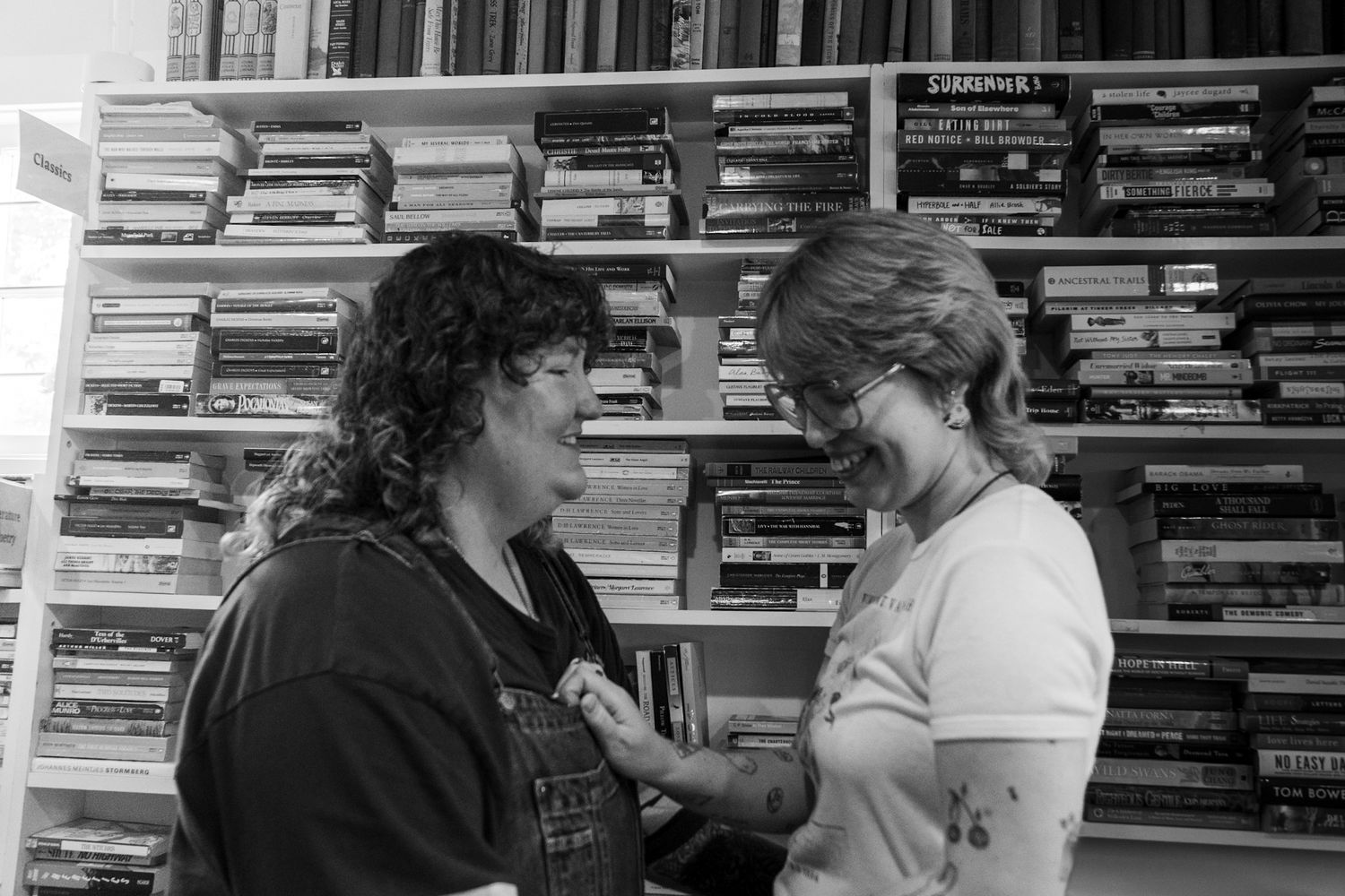 Two women engaged in conversation in front of shelves filled with books and VHS tapes in a black and white photo.