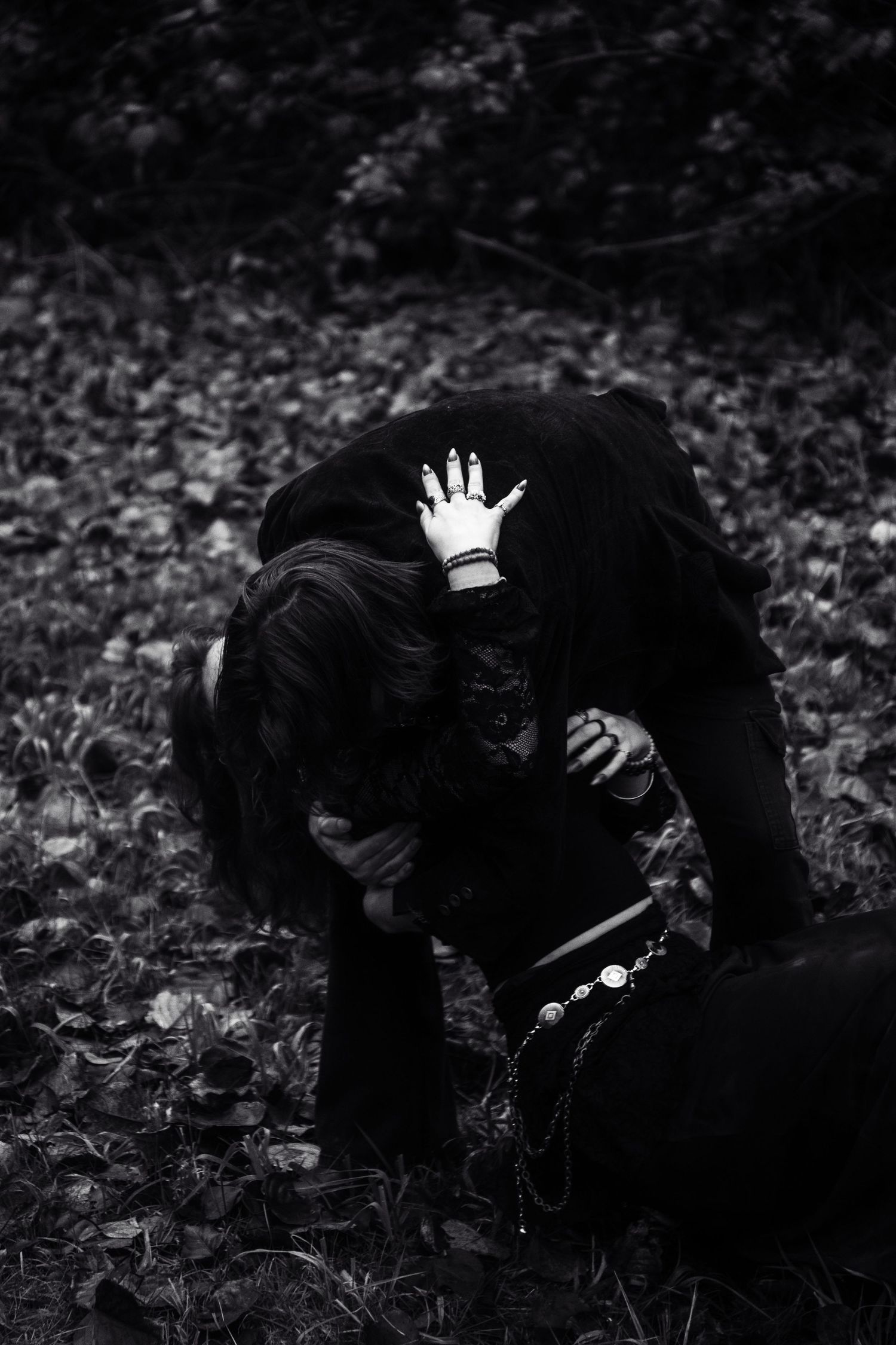 Black and white image of two people in black embracing on forest floor.