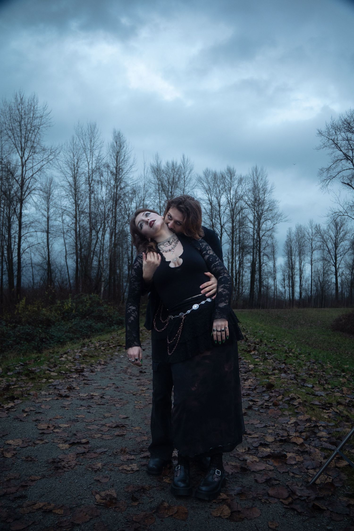 Gothic couple in black embrace on a leaf-covered path surrounded by bare autumn trees.