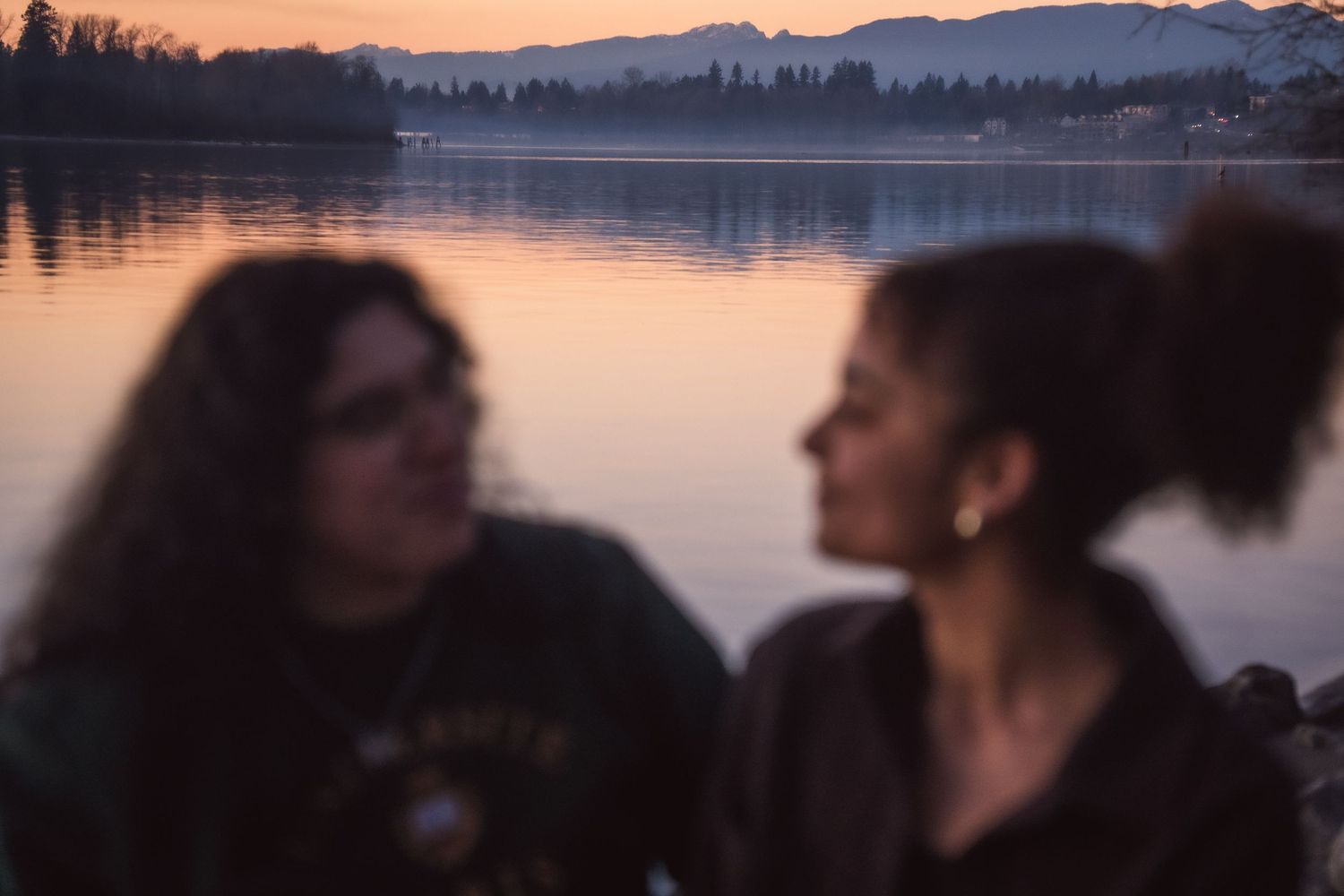 Two women in soft focus at lakeside during golden hour sunset with mountains.