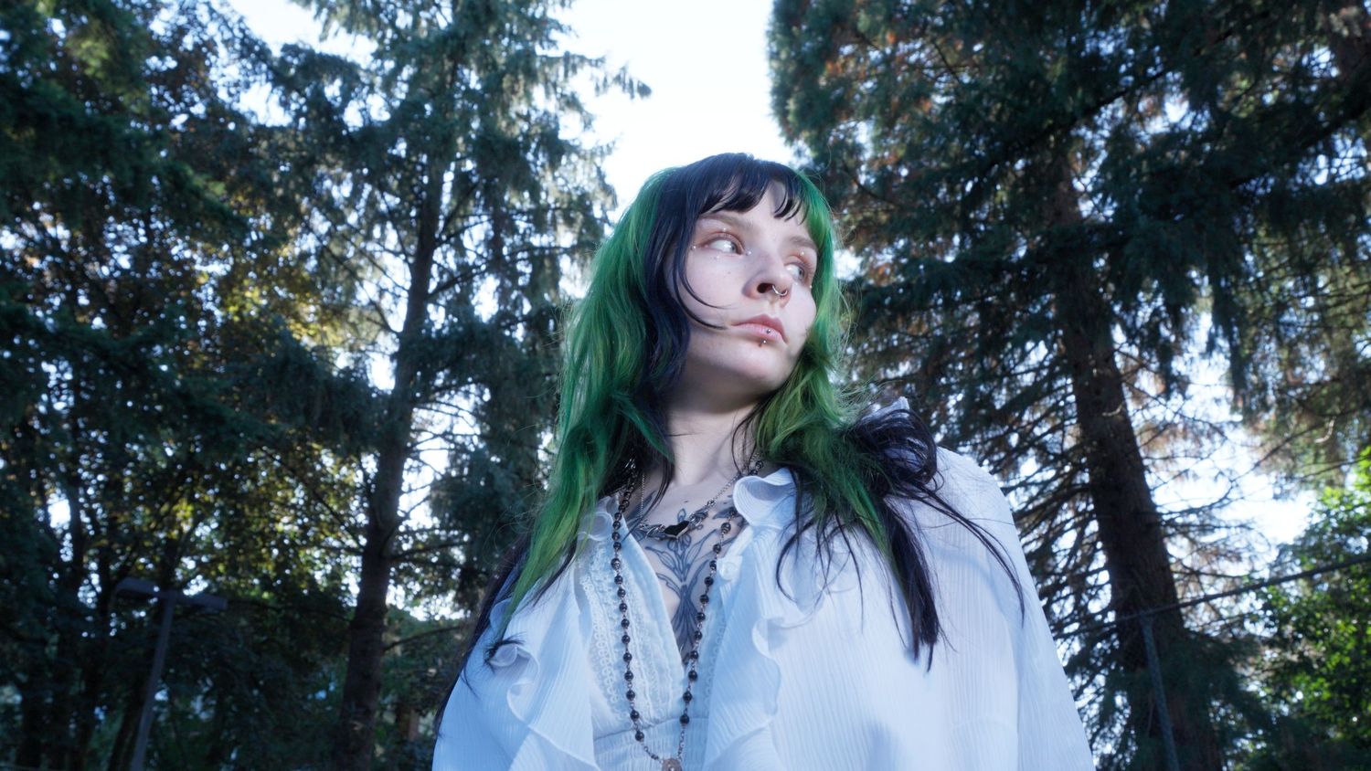 Woman with green hair in white blouse looks up among tall trees outdoors.