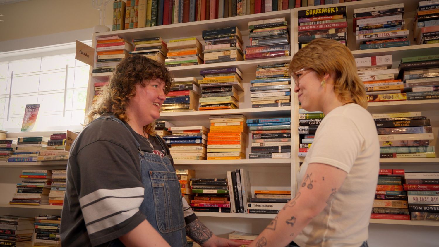 Two young women smiling and holding hands facing each other in a cozy bookstore.