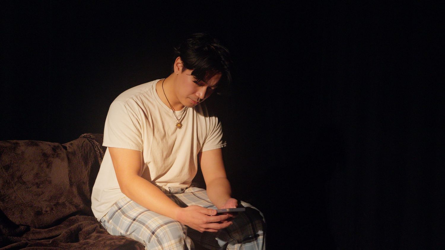 Young Asian man sits alone on a bed in a dark room, looking down at his phone.