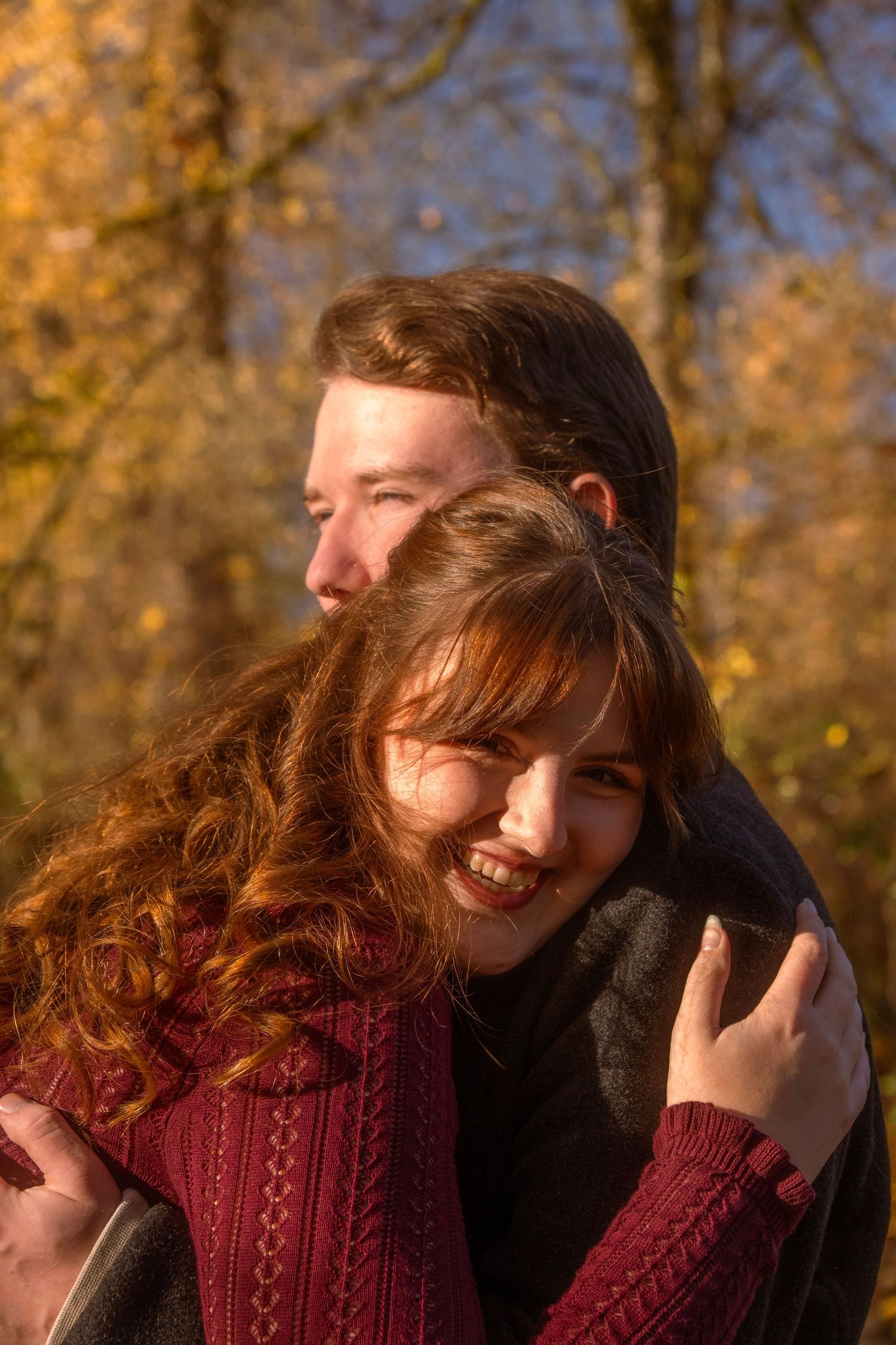 Young couple embracing outdoors in autumn forest, woman smiling with curly red hair, golden fall foliage background.