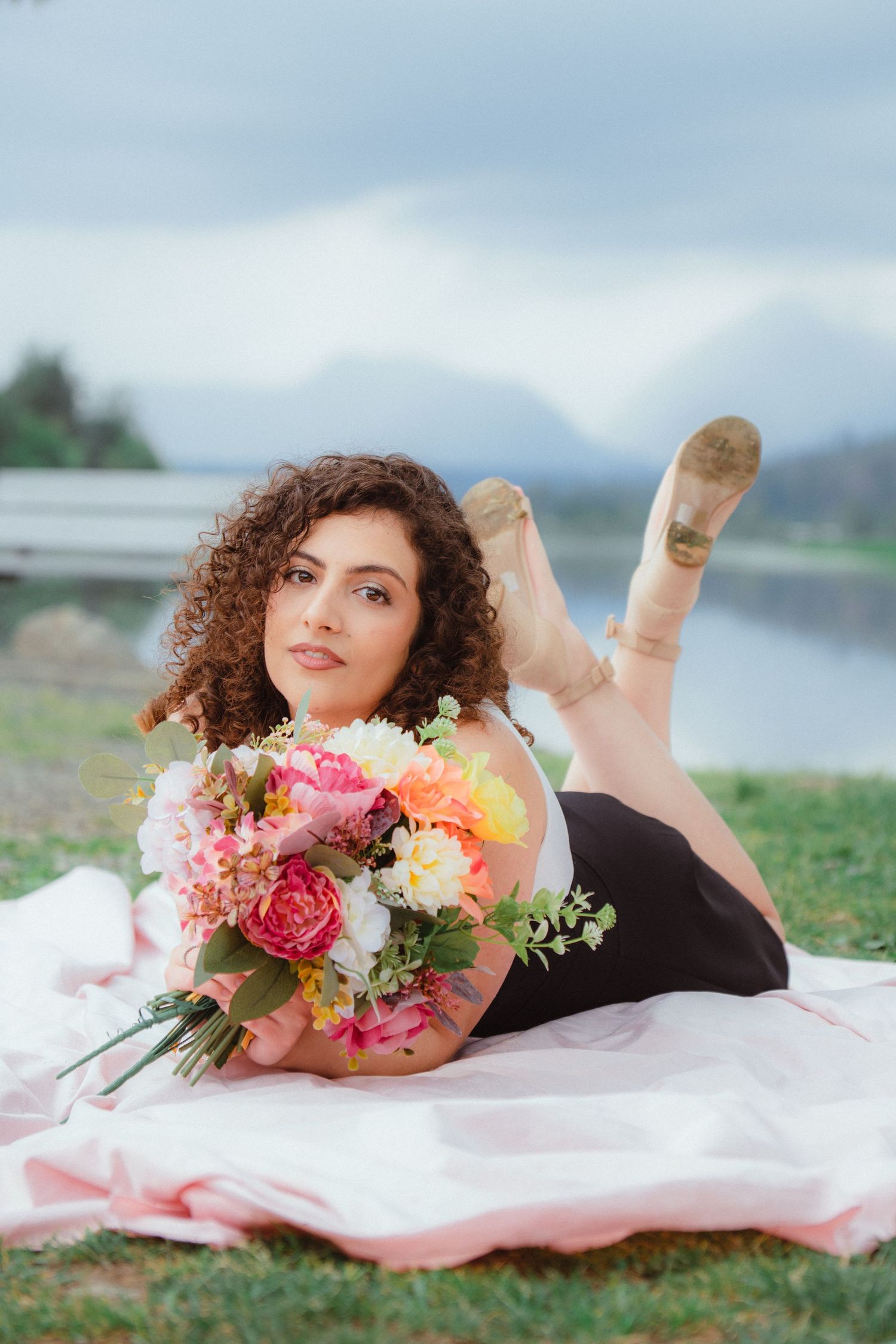 Curly-haired woman in black dress lying on blanket holding vibrant floral bouquet near mountain lake.