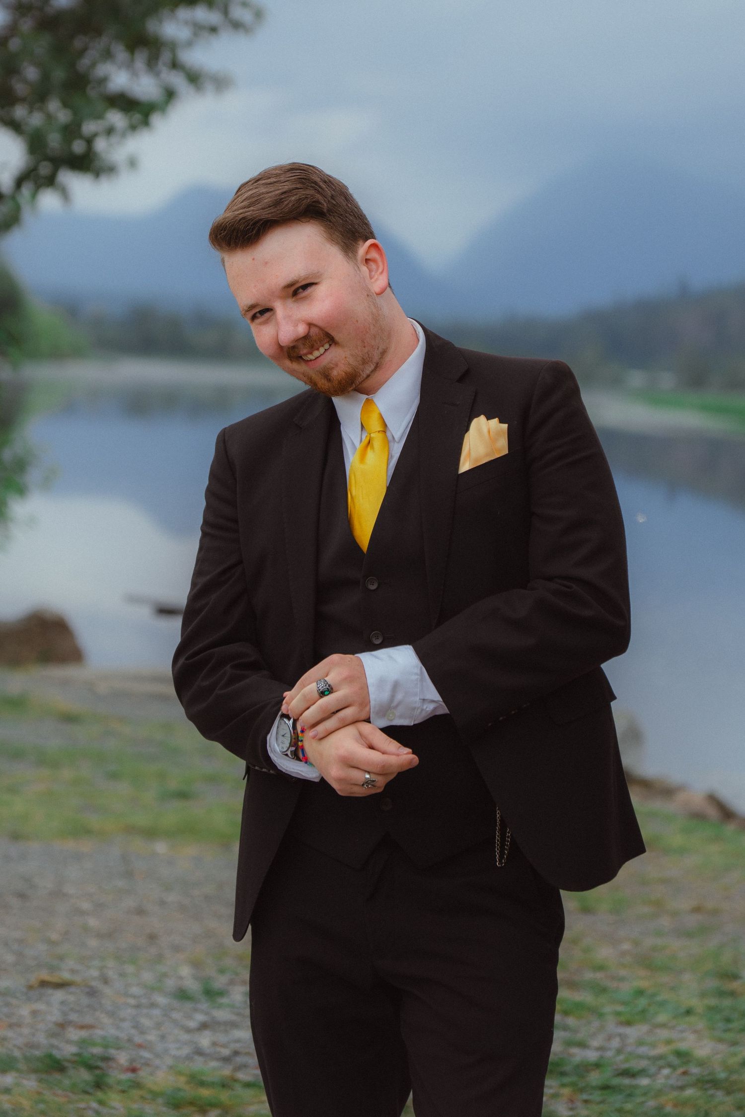 Groom in black suit adjusts cufflinks while smiling by a misty mountain lake.