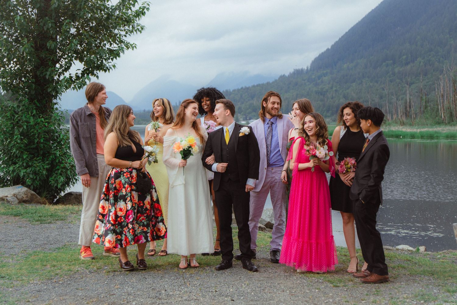 Bride and groom with wedding guests in vibrant attire posing by a mountain lake backdrop.