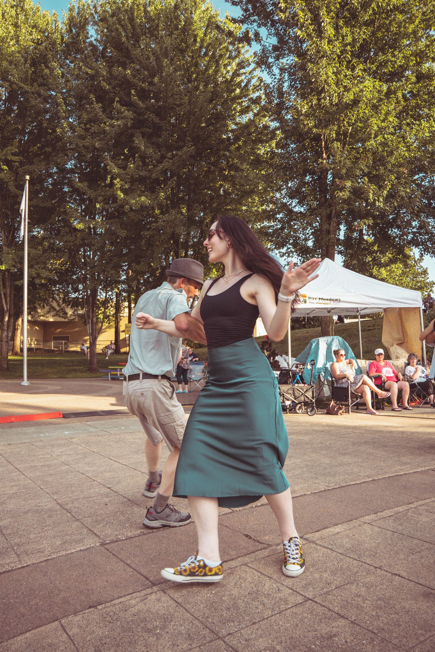 Woman in teal skirt and sneakers dancing outdoors at a summer festival with trees and a white tent in the background.