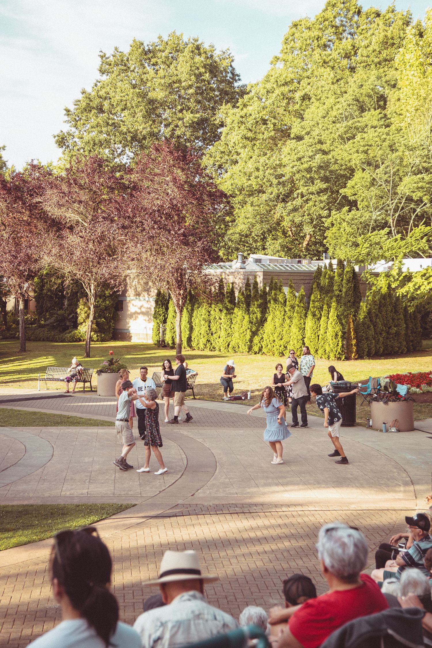 People dancing outdoors in a sunny park plaza, surrounded by lush green trees and spectators enjoying the event.