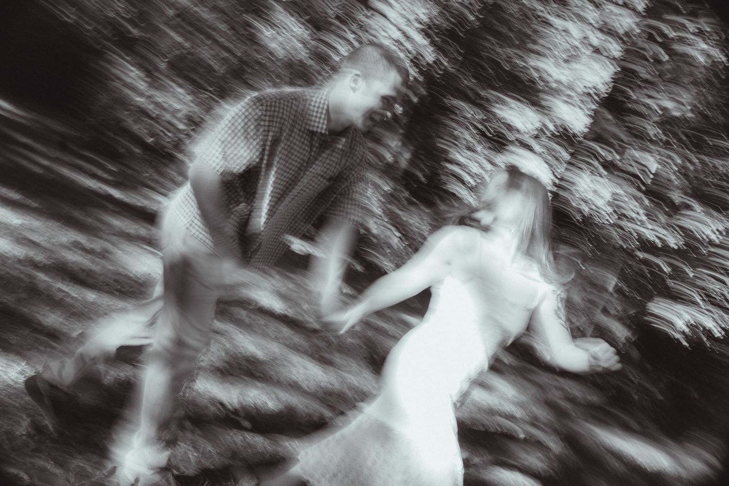 Blurred black and white photo of a couple dancing outdoors among trees, capturing motion and romance.
