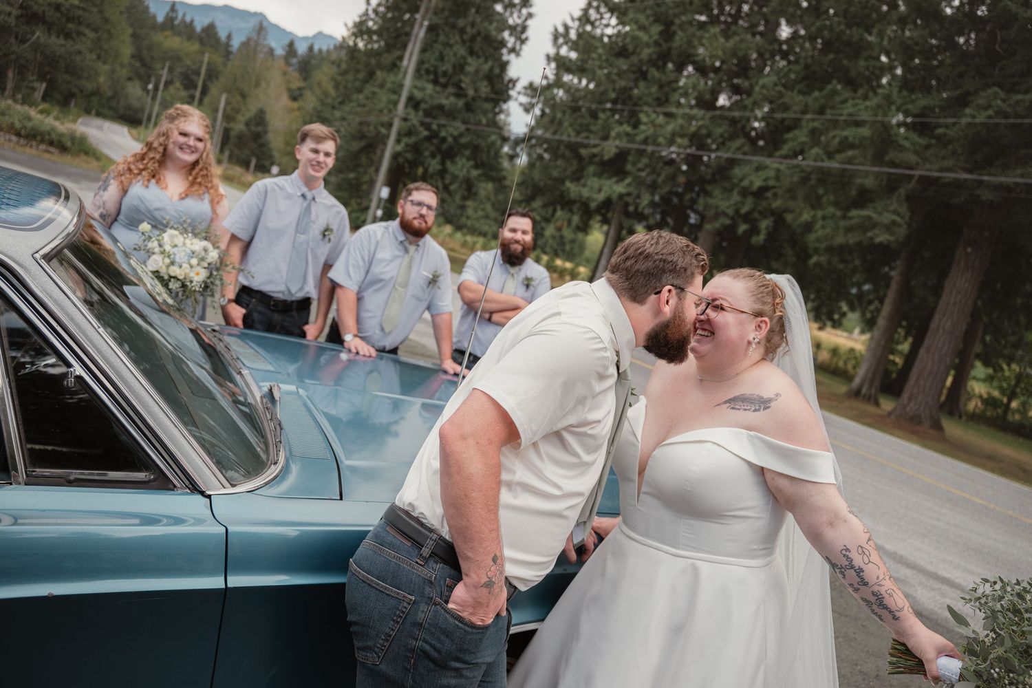 Bride and groom share a kiss beside a vintage teal car while wedding party poses in background outdoors.