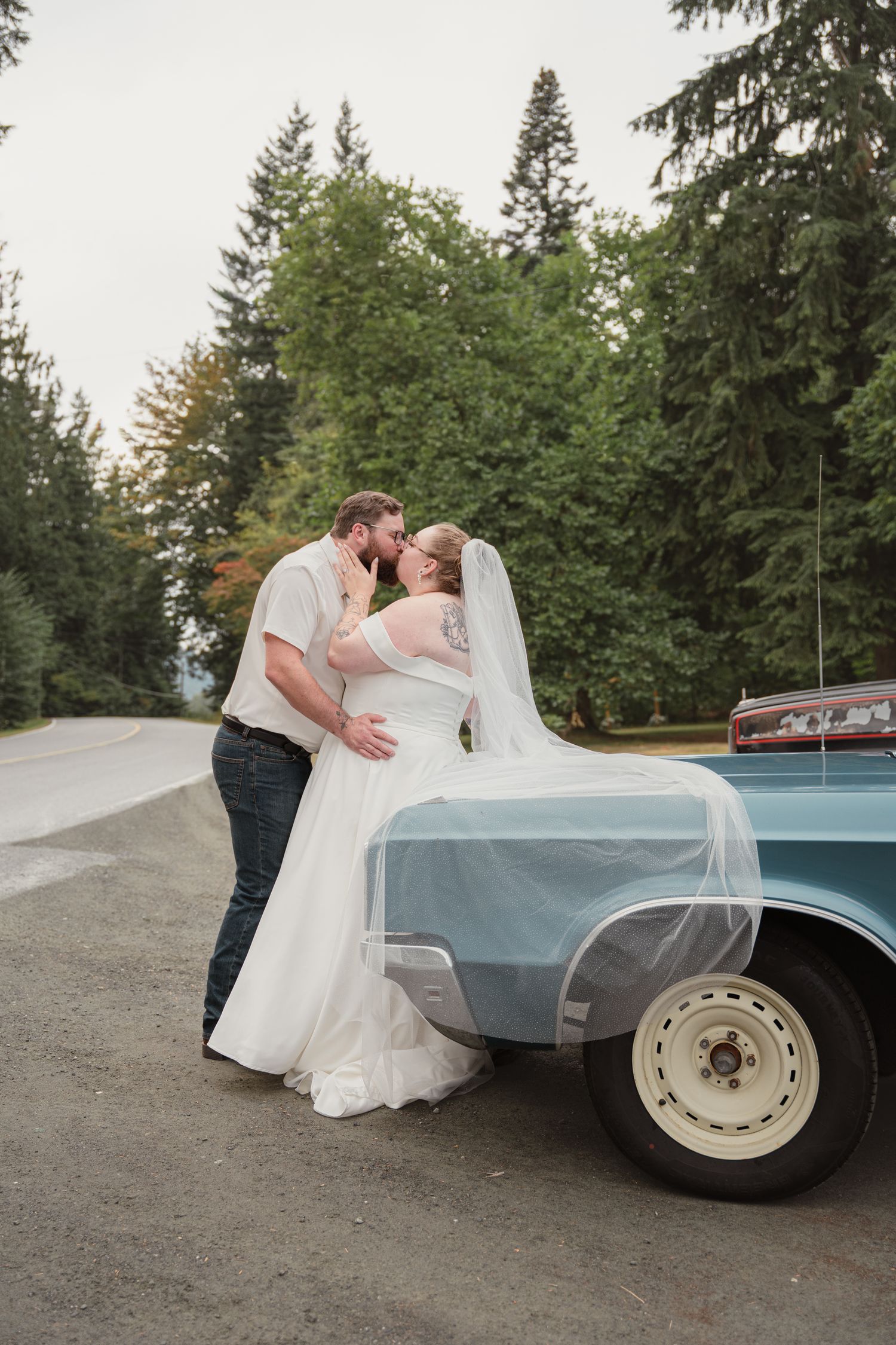 Bride and groom kissing beside a vintage light blue truck, surrounded by tall evergreen trees on their wedding day.