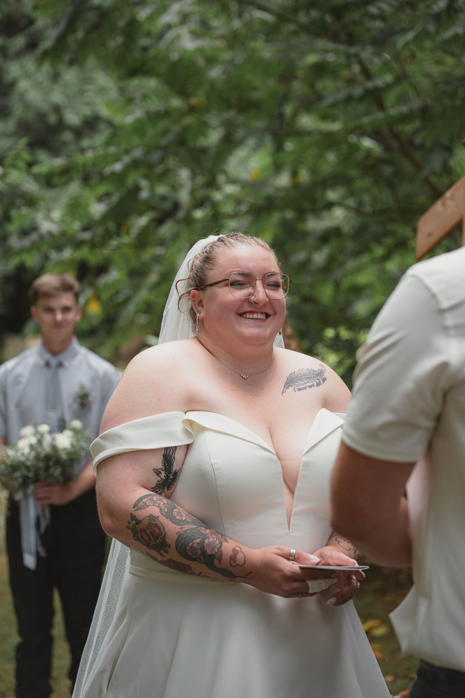 Tattooed bride in off-shoulder white gown and veil smiling radiantly during outdoor wedding ceremony.