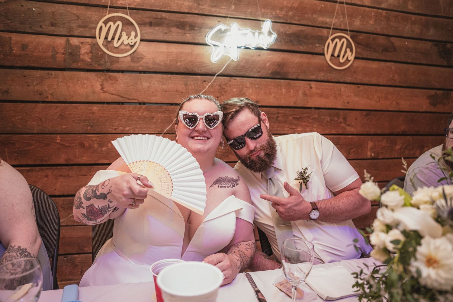 Newlywed couple in heart sunglasses celebrating at wedding reception with neon Mr and Mrs sign backdrop.