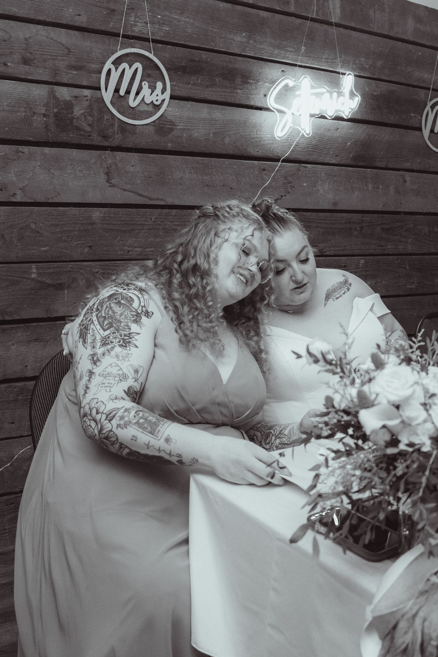Two brides share a tender moment at signing table beneath Mrs neon sign in black and white wedding portrait.
