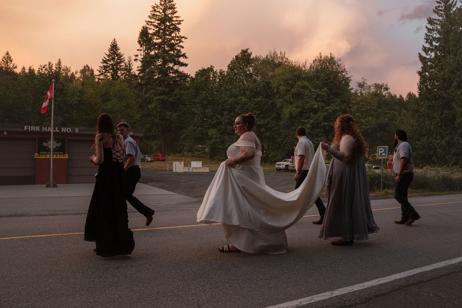 Bride in white gown with wedding party walking across parking lot at sunset with dramatic orange sky backdrop.