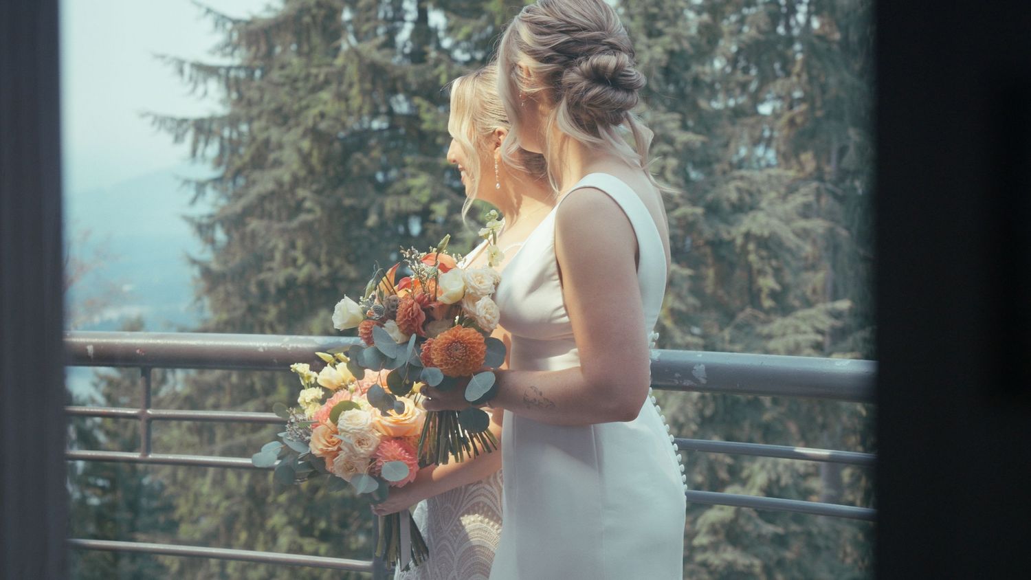 Two brides in white dresses holding colorful bouquets on a balcony surrounded by lush forest trees.