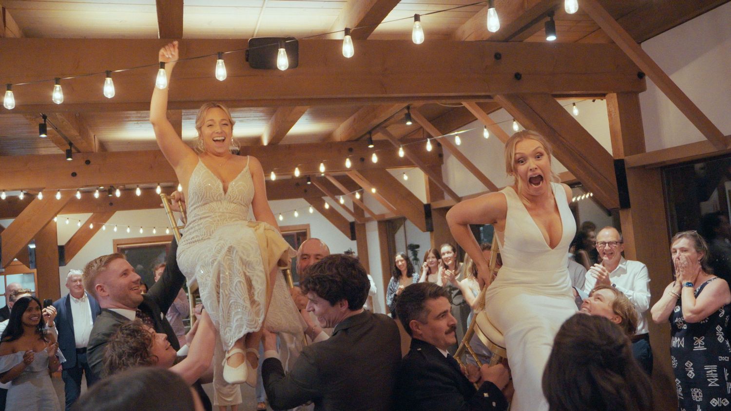 Two brides in white gowns are lifted by guests during a joyful wedding reception celebration.