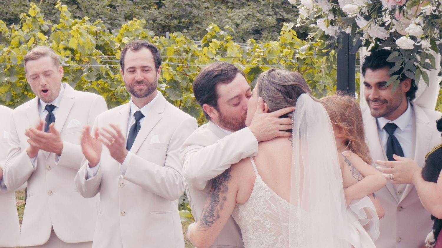 Bride and groom share first kiss at outdoor vineyard wedding ceremony surrounded by groomsmen in cream suits.