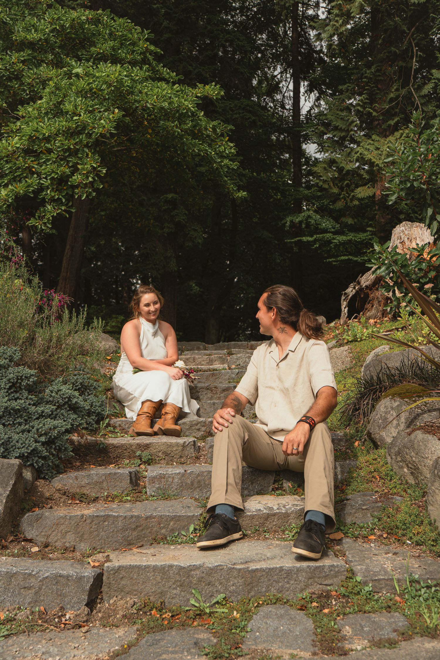 Bride and groom sitting casually on stone steps in a lush forested garden setting.