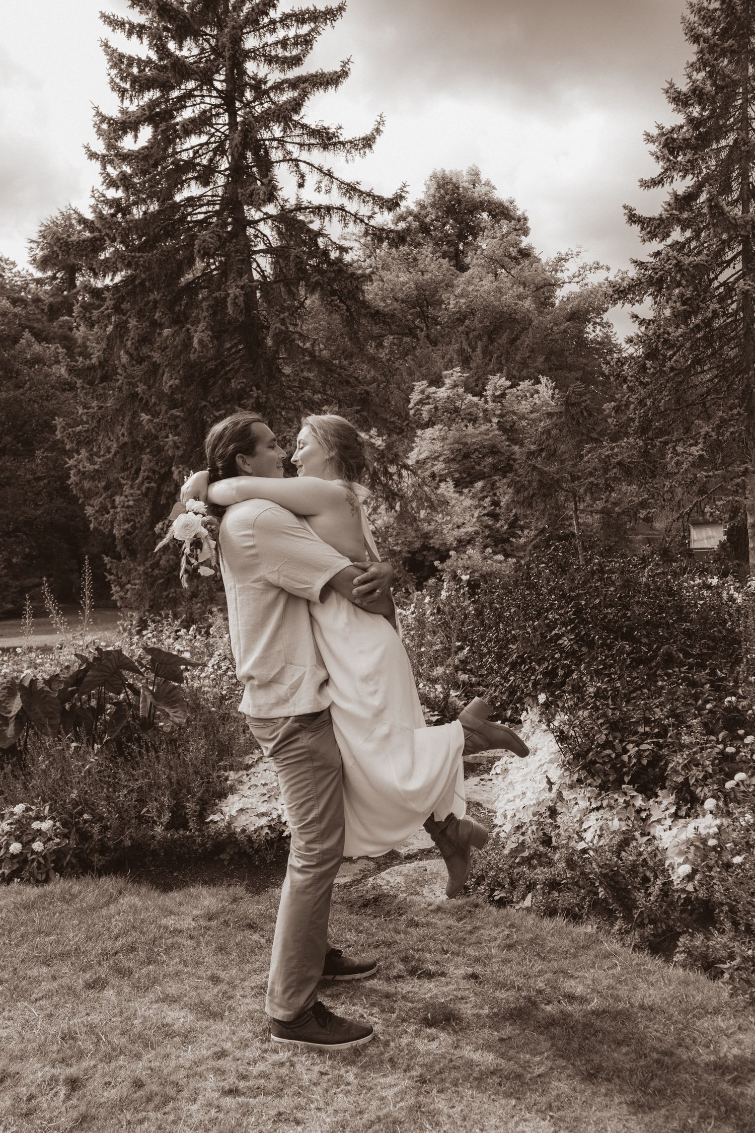 Groom lifts bride in a joyful embrace in a lush garden in this romantic sepia-toned wedding photo.