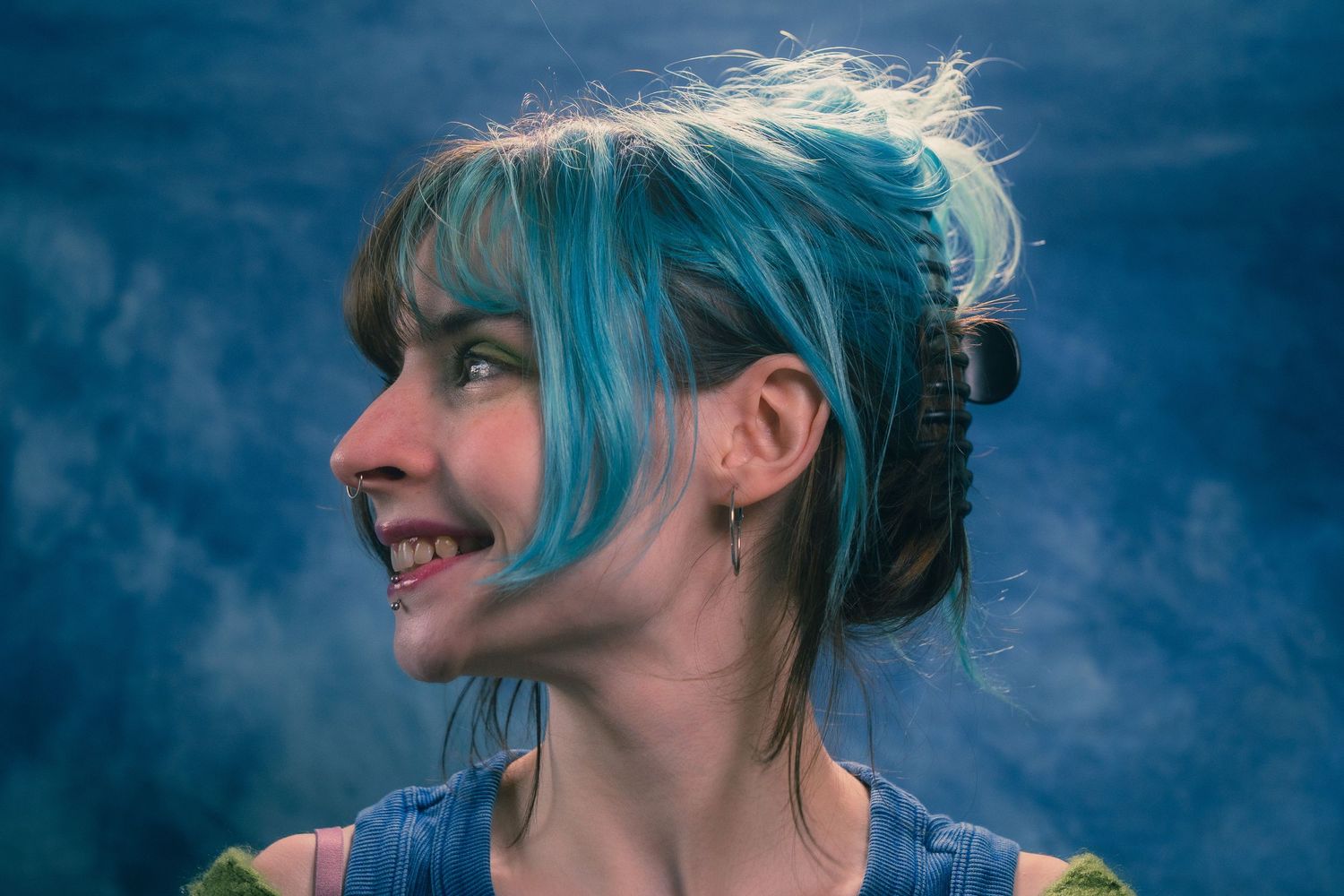 Side profile of smiling woman with blue braided updo hair and hoop earring in blue studio setting.
