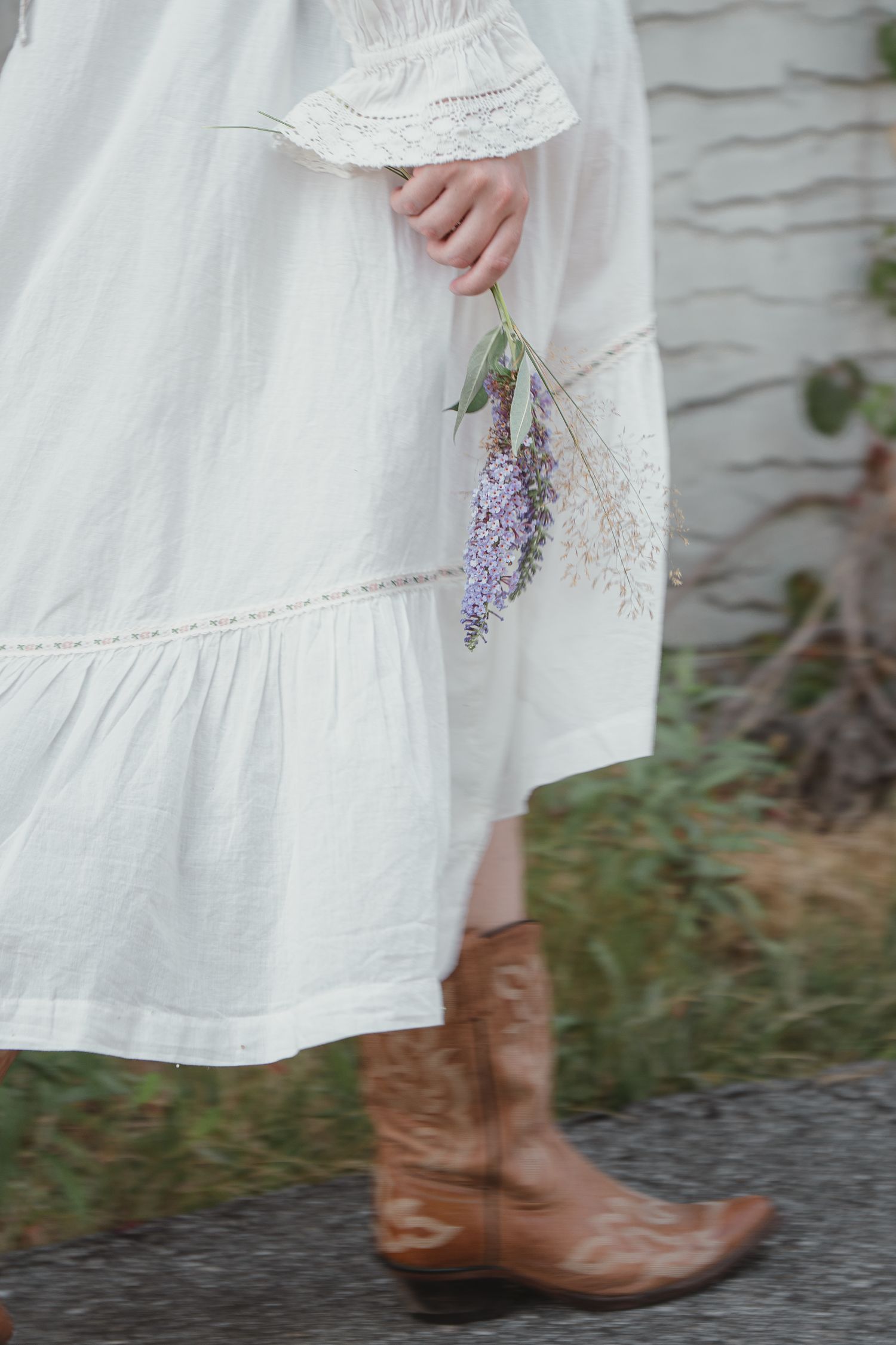Detail shot of white linen tiered dress with lace trim, brown cowboy boots, and purple lavender stems.