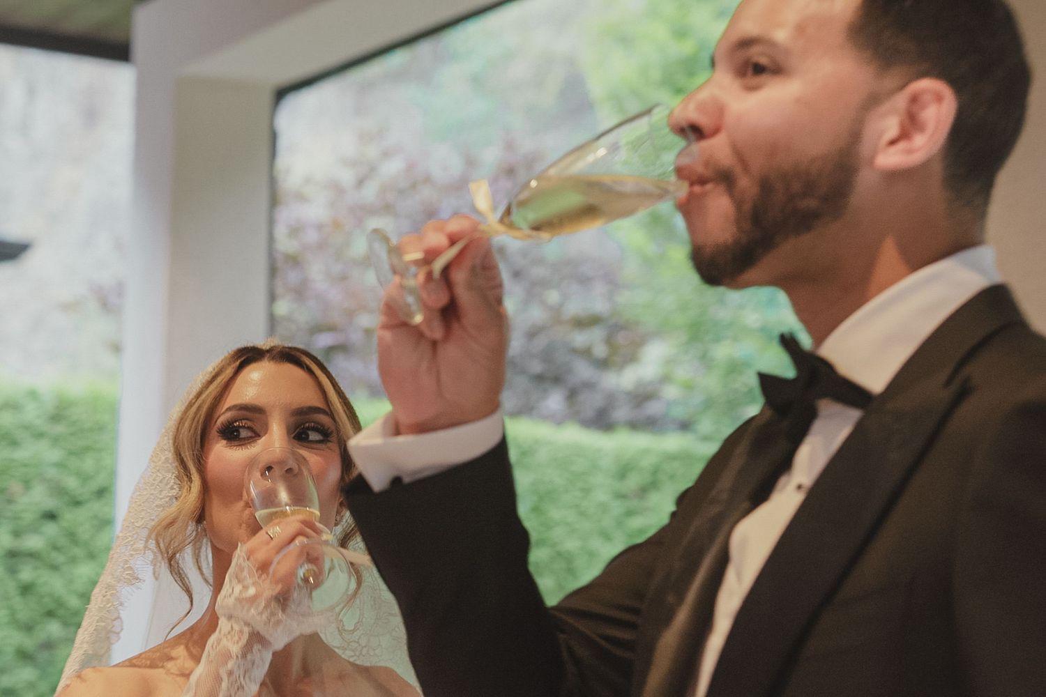 Bride and groom drinking champagne toast at wedding reception in elegant venue with garden views.
