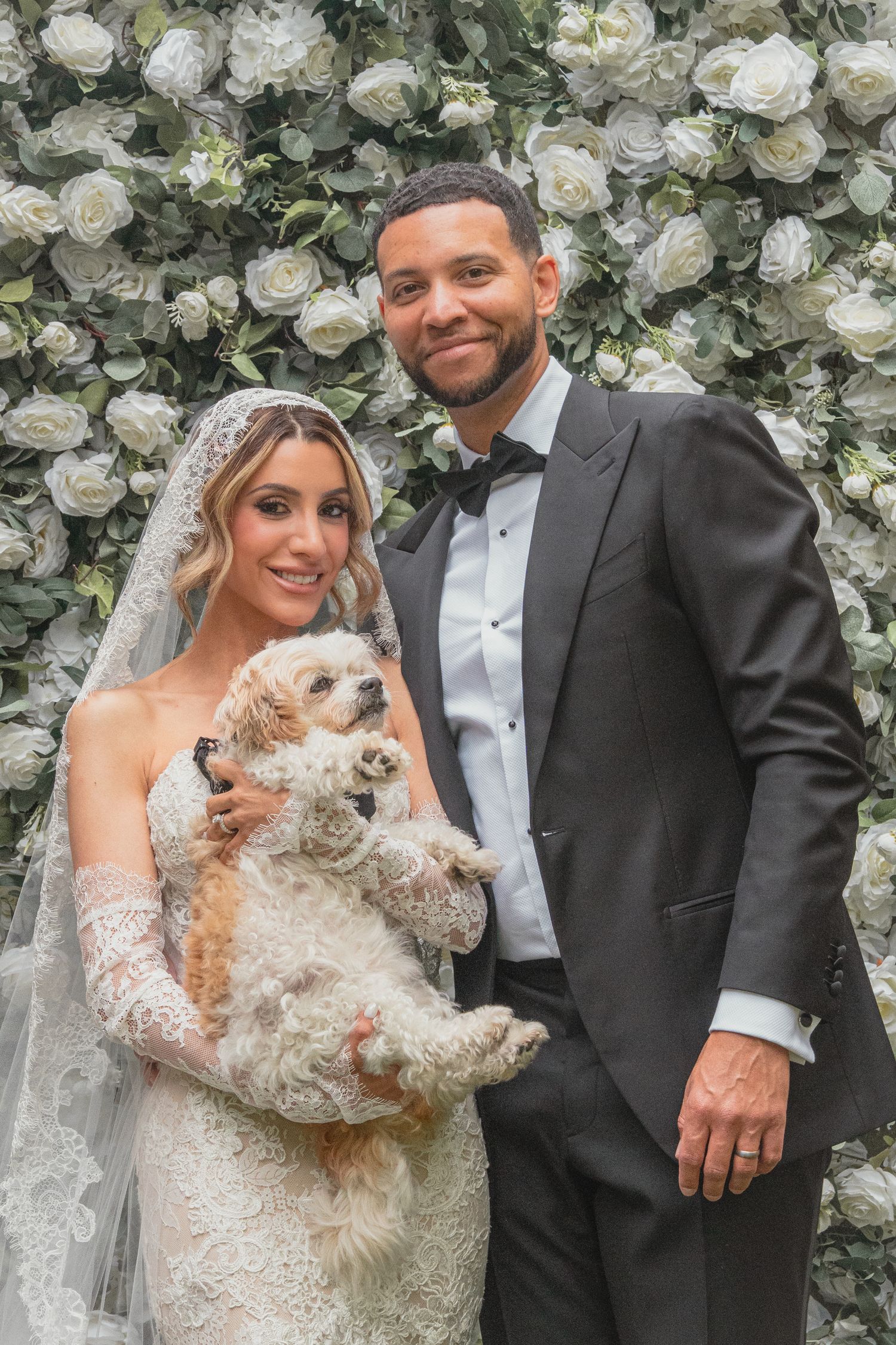 Bride and groom posing with a small fluffy dog in front of a white floral wall on their wedding day.