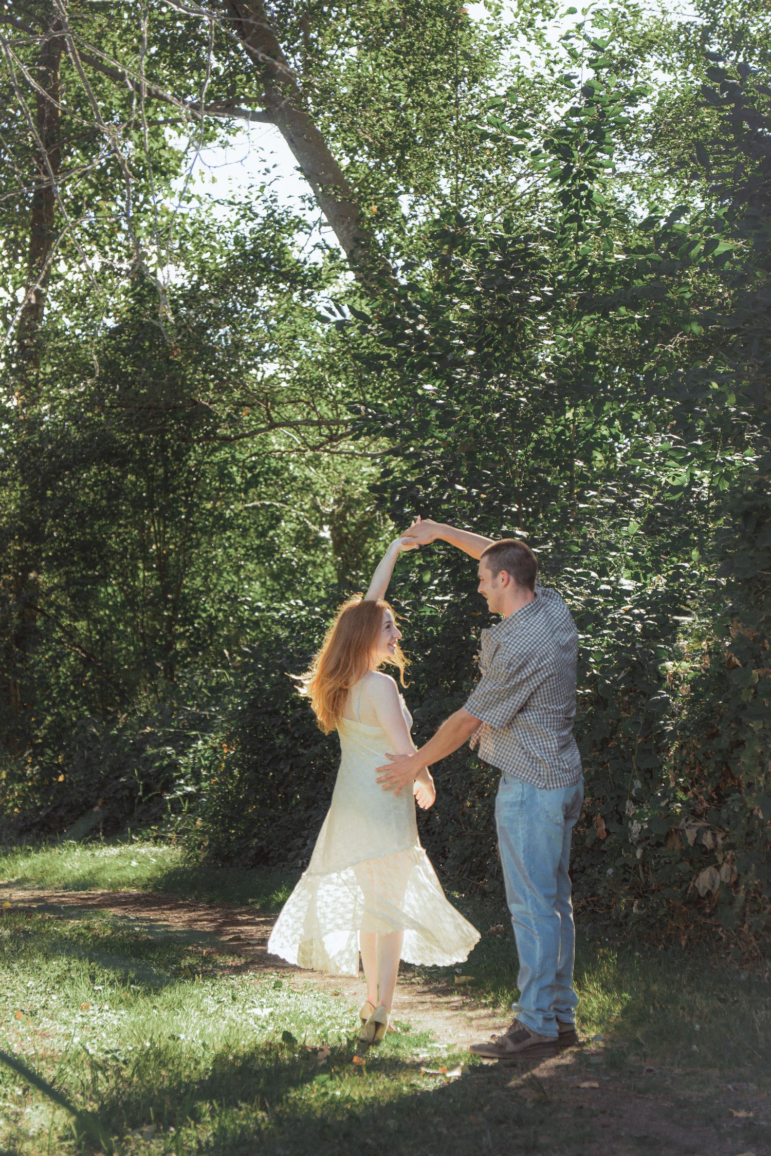 Couple dancing outdoors in sunlit forest, woman in white dress twirling, man in striped shirt and jeans.