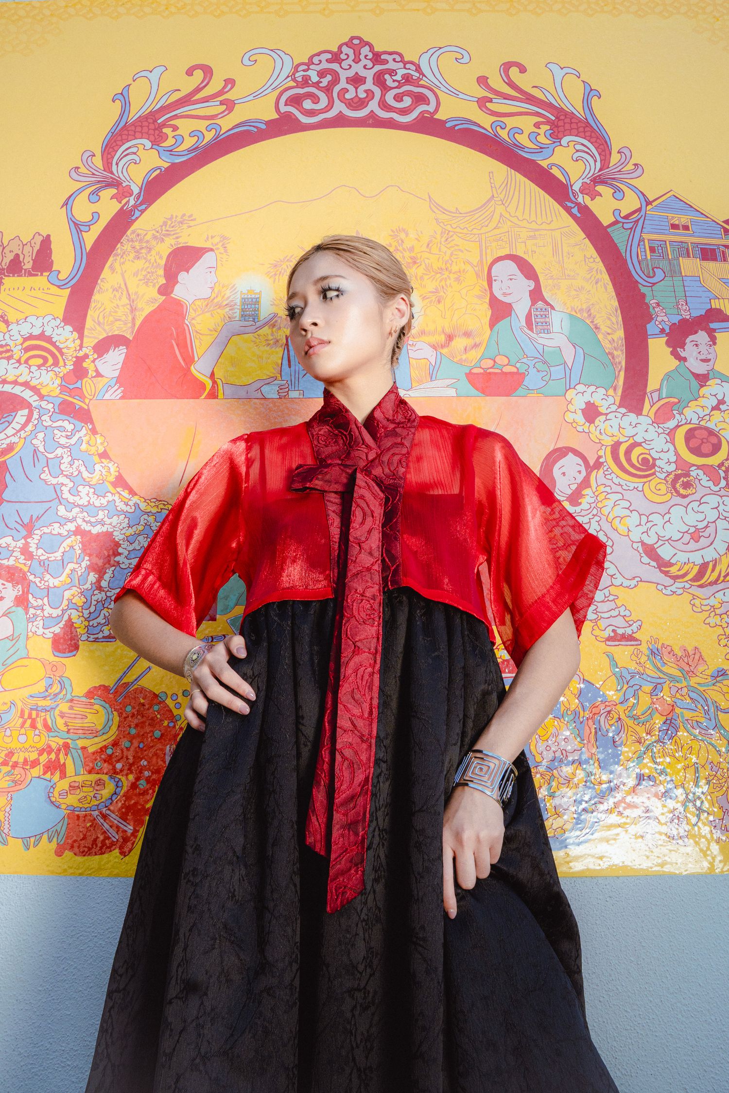Woman in red satin cape and black skirt poses before colorful Chinese mural art.