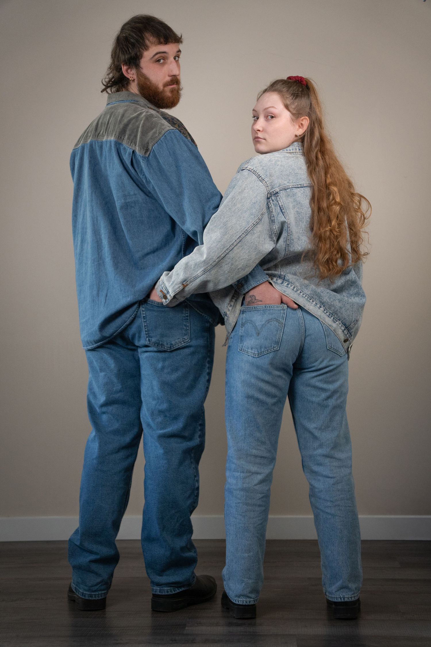A couple in coordinating denim outfits stand back to back, glancing over their shoulders at the camera.