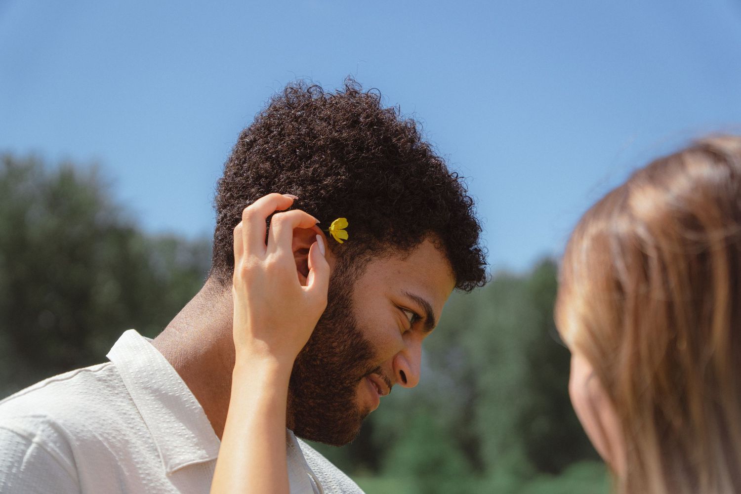 A woman gently placing a small yellow flower behind a curly-haired mans ear against a clear blue sky.