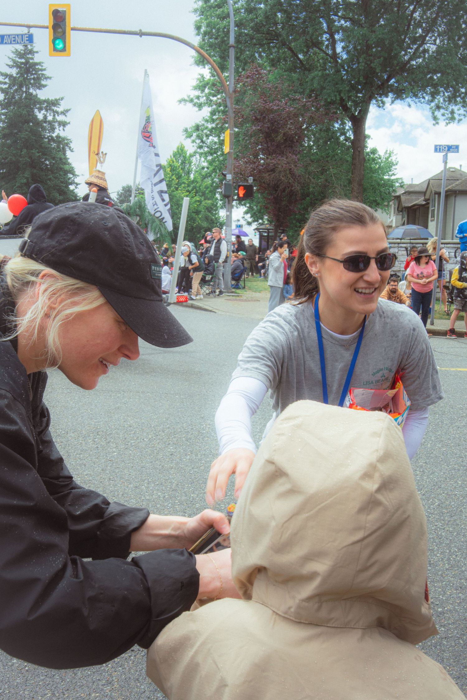 Two smiling women interact with a seated parade participant on a busy street during a community parade.