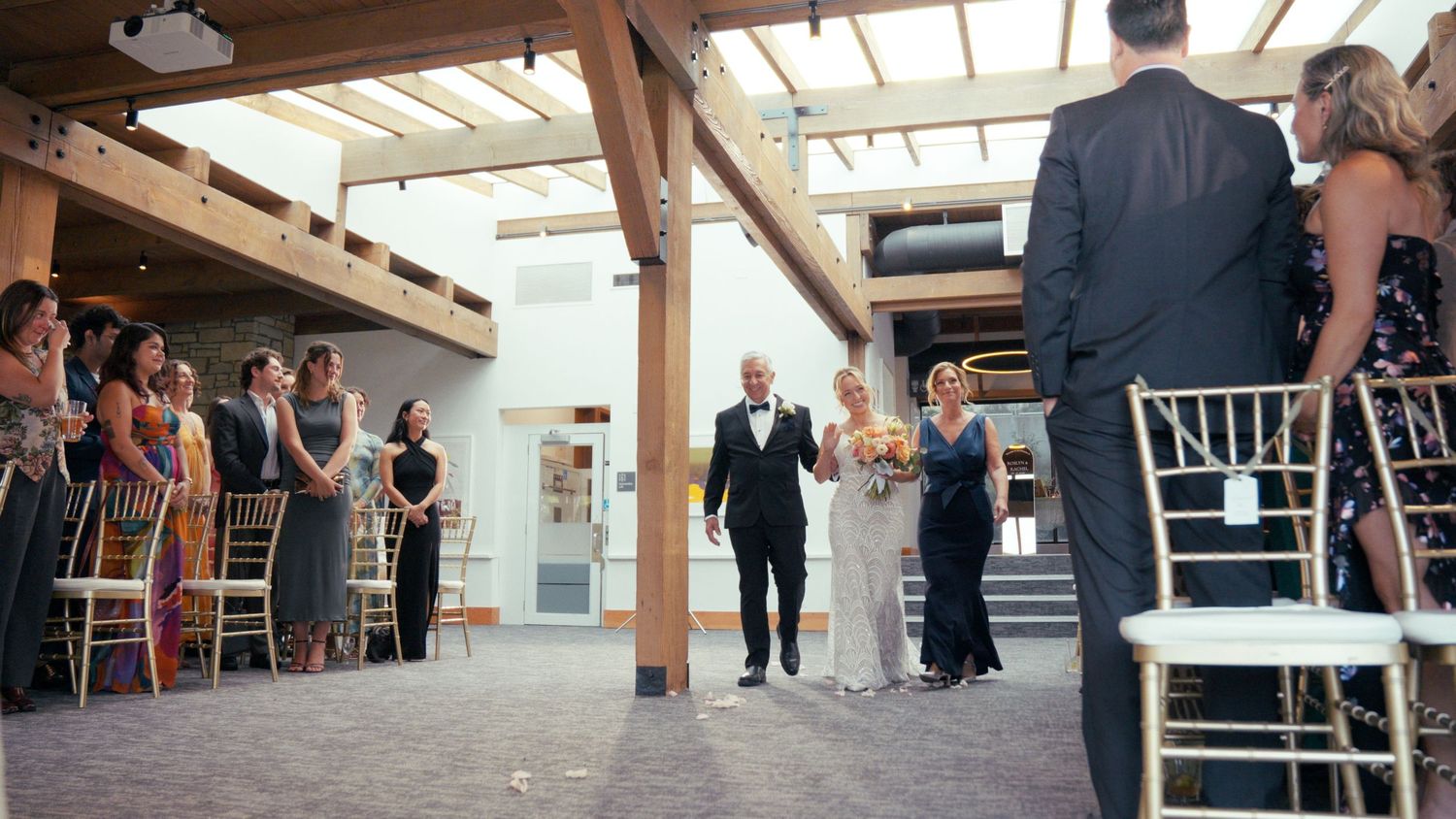 Bride walking down the aisle with escort at indoor wedding ceremony with seated guests.