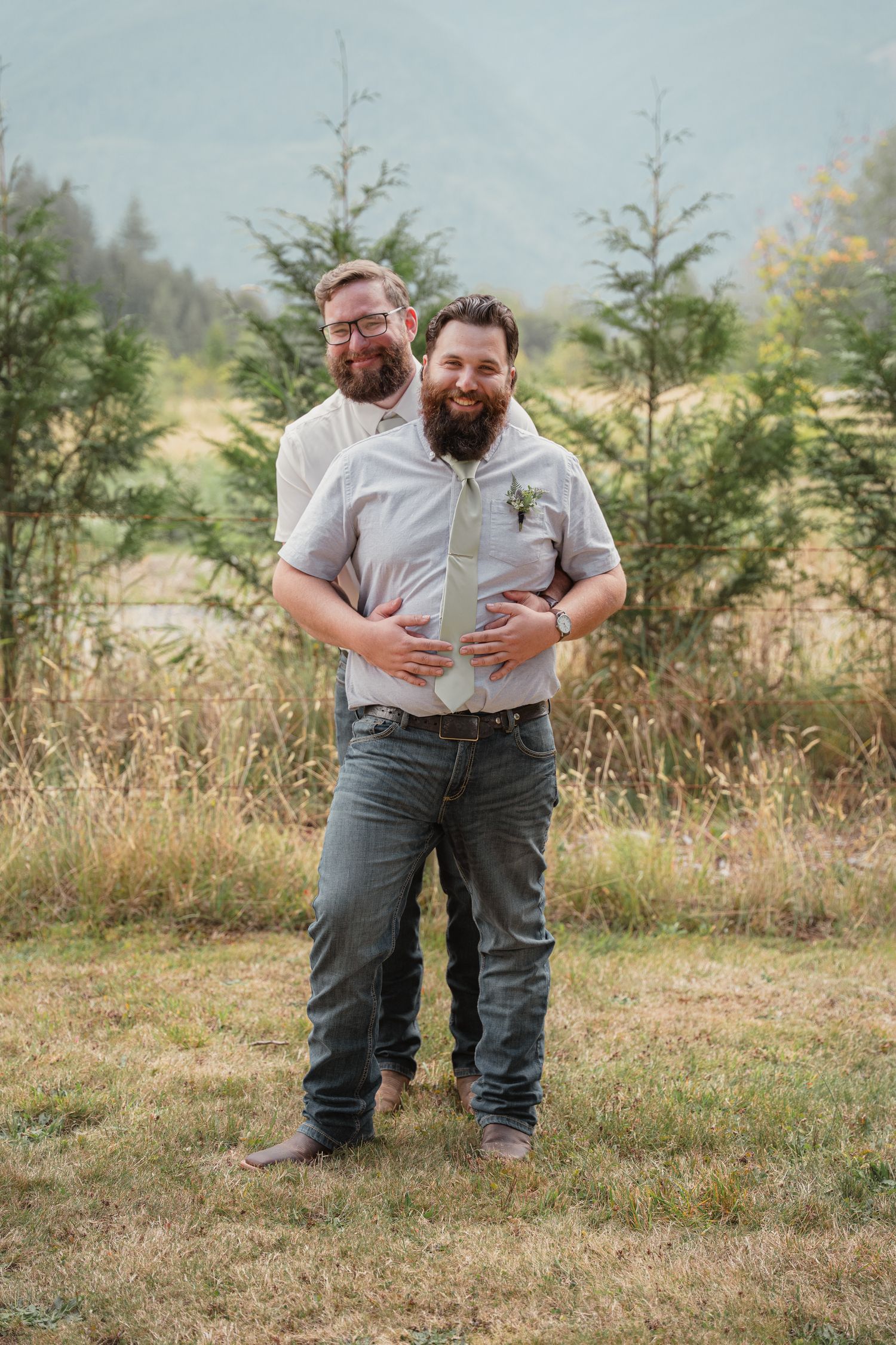 Two bearded grooms share a playful embrace outdoors with mountain scenery during their wedding day.