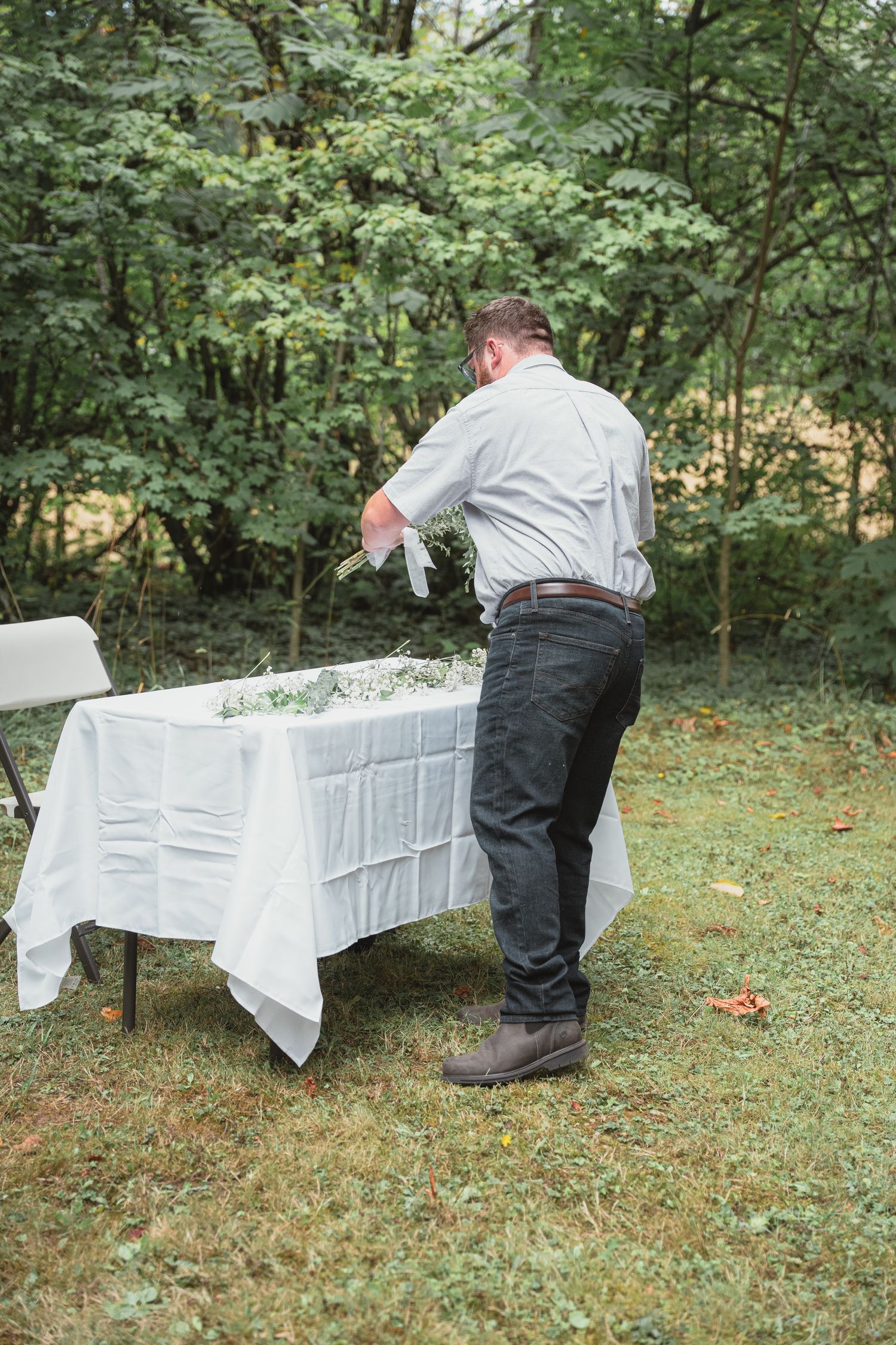 Man arranging white floral centerpiece on outdoor table with white tablecloth in garden setting.