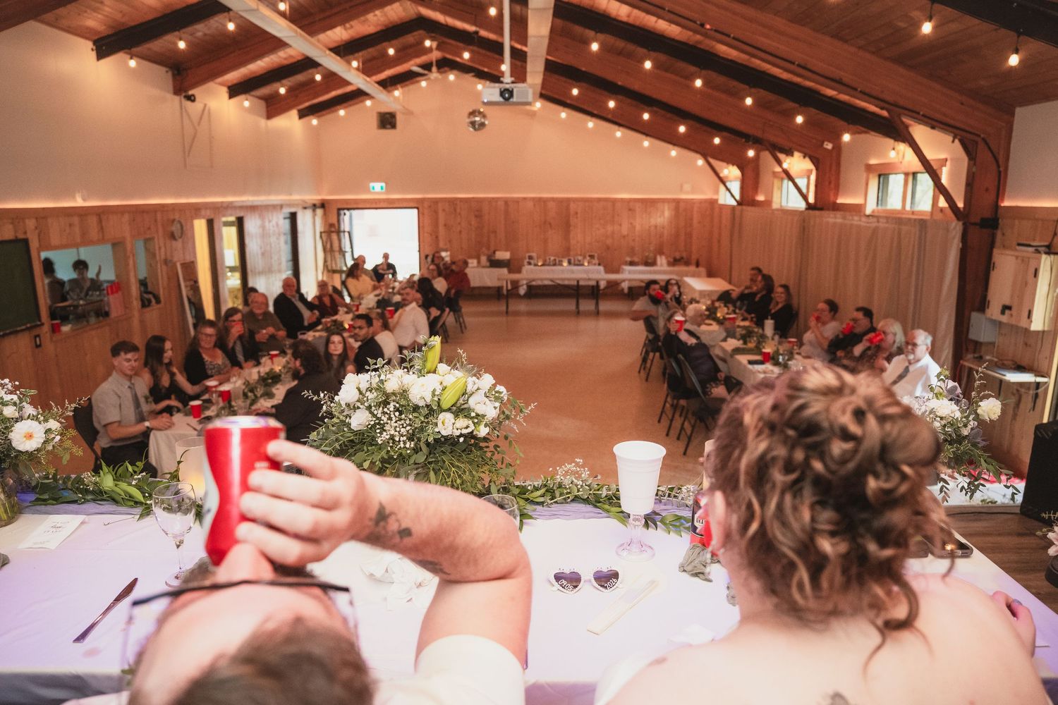 Overhead view of wedding reception in rustic barn venue with guests seated at decorated tables with floral centerpieces.