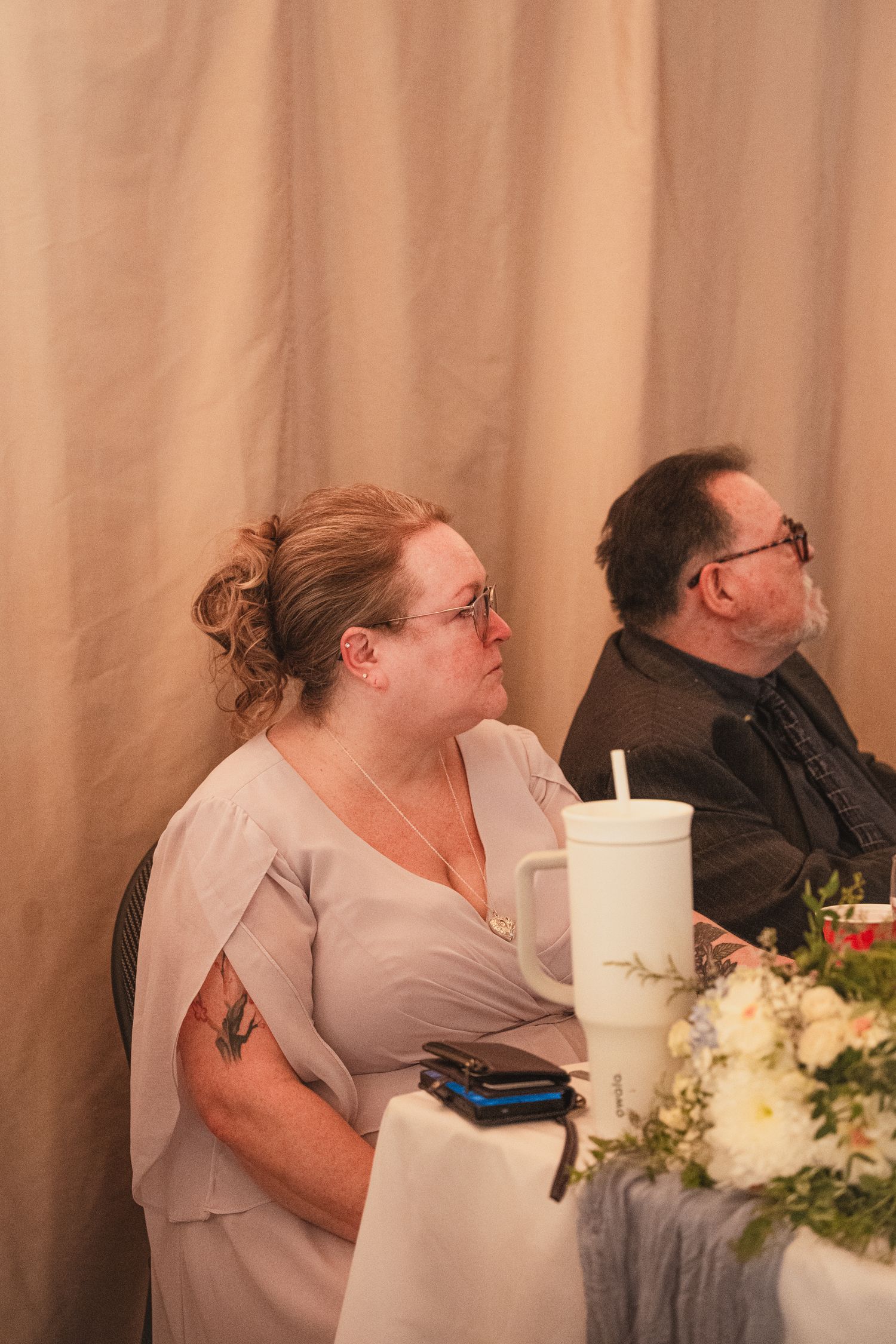 Woman in gray dress with tattoo sits at wedding reception table beside man in dark jacket.