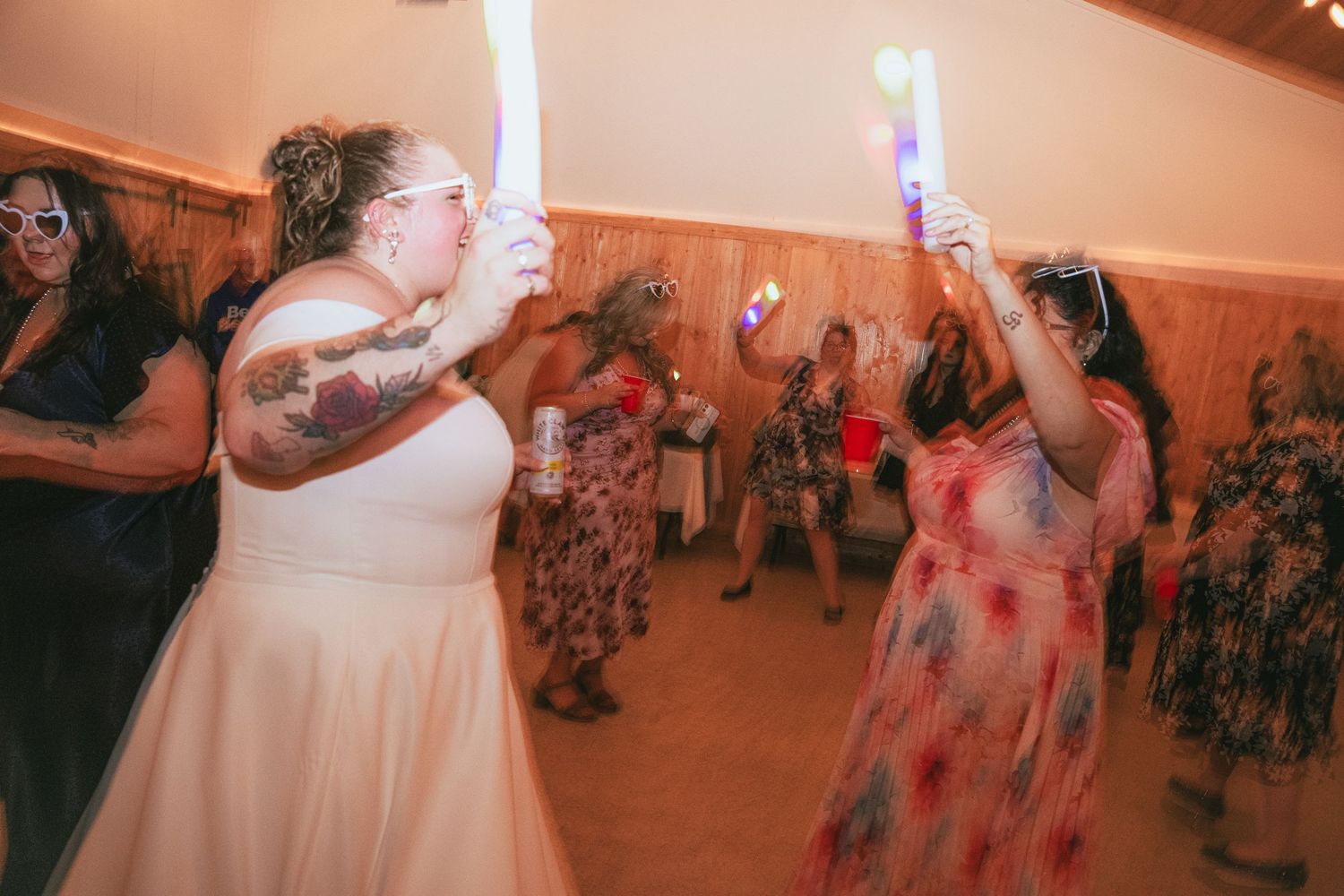 Bride in white dress dancing with guests holding glow sticks at a lively wedding reception party.