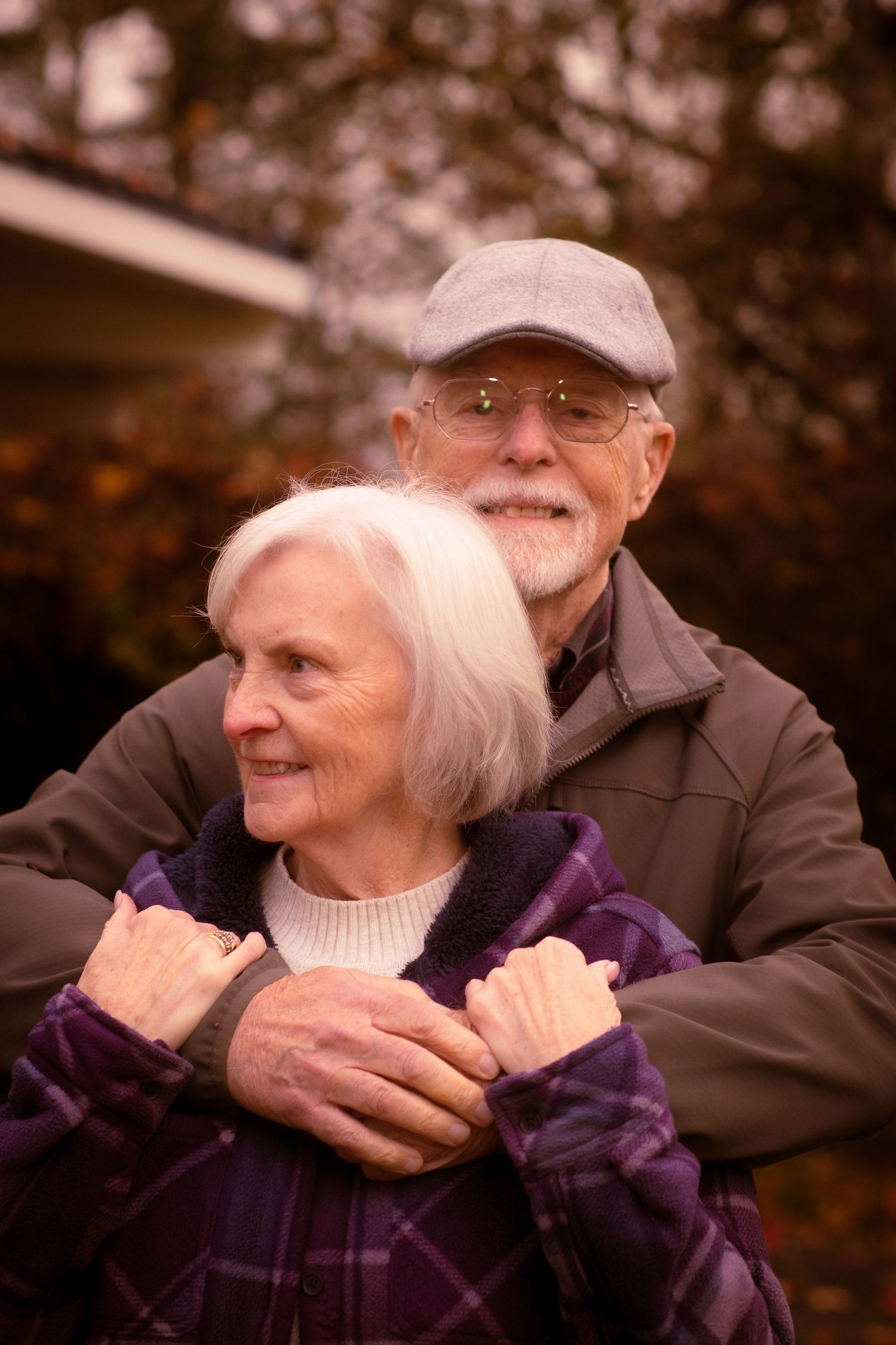Elderly man hugging woman from behind, both smiling in a fall garden setting.