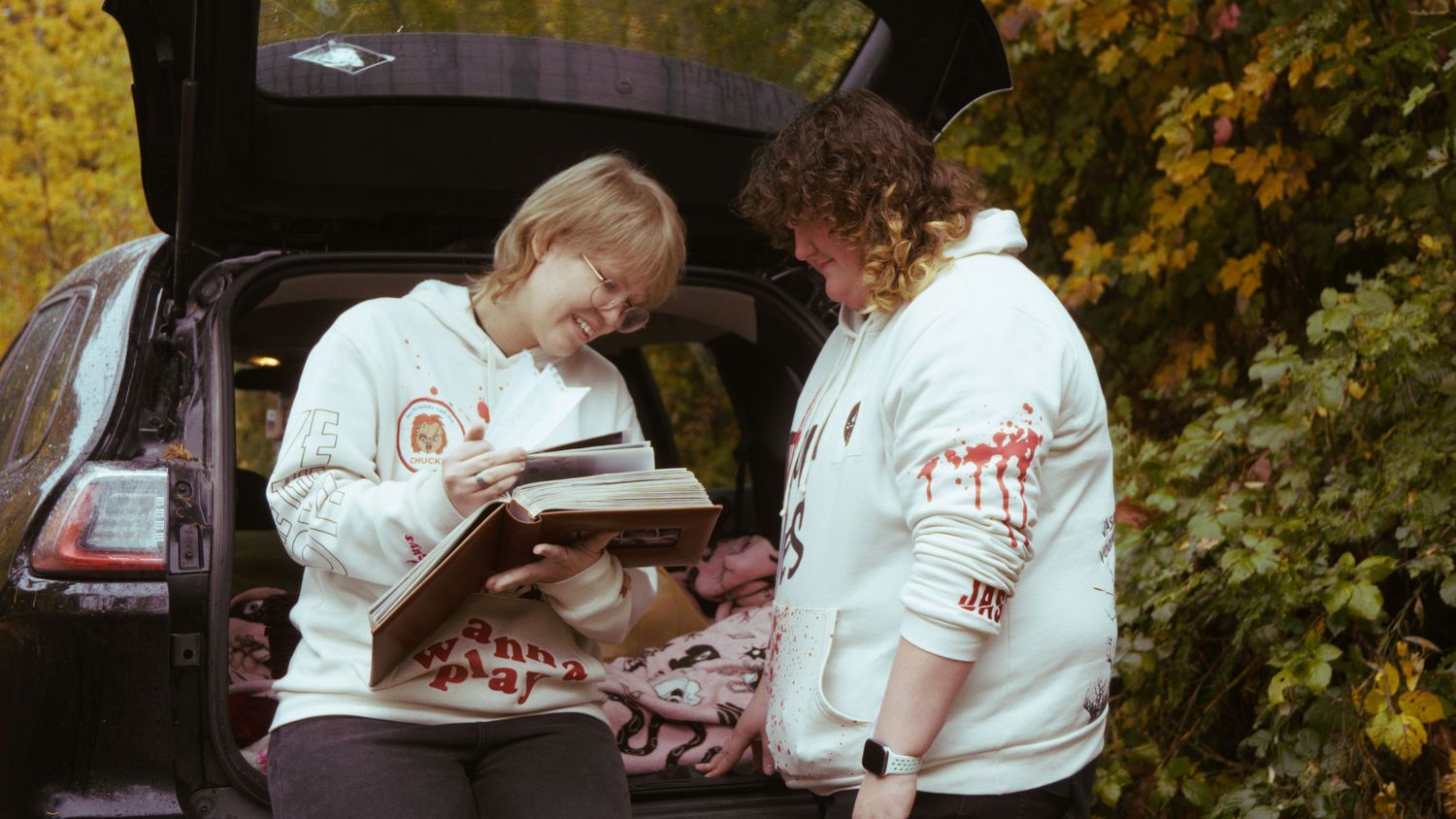 Two people in white hoodies look at a photo album together at the open trunk of an SUV.